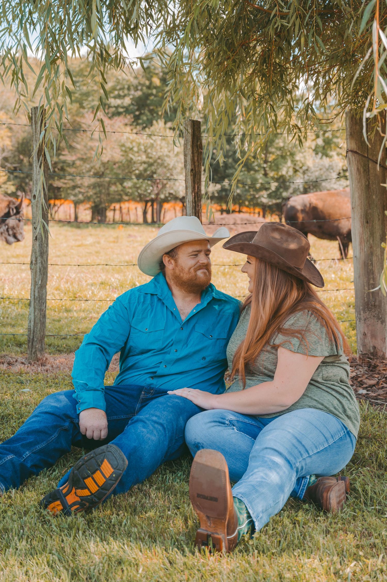 A couple in cowboy hats relaxing together on grass under willow trees.