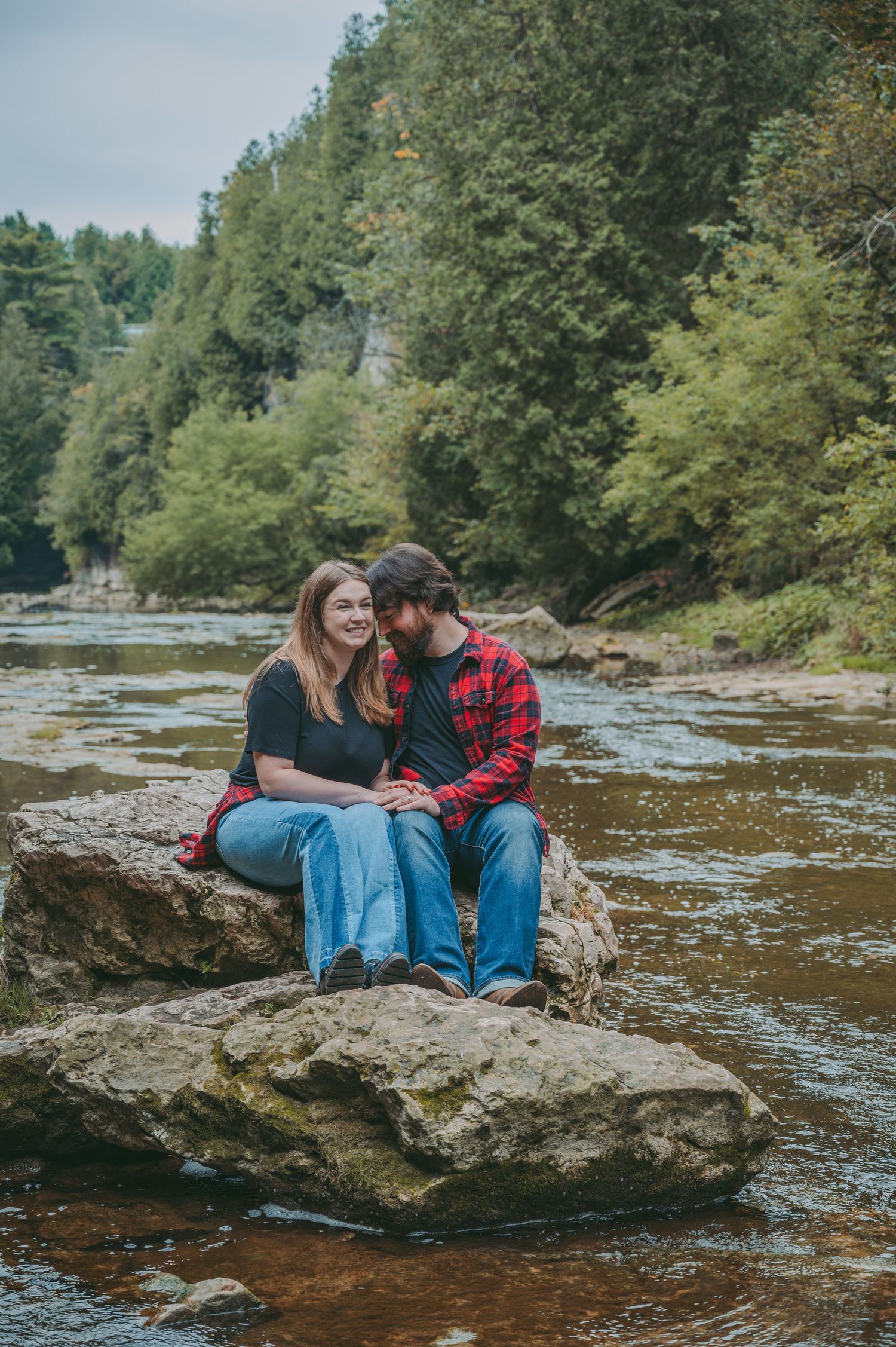 Happy couple sharing tender moment while sitting on rocky riverbank surrounded by forest.