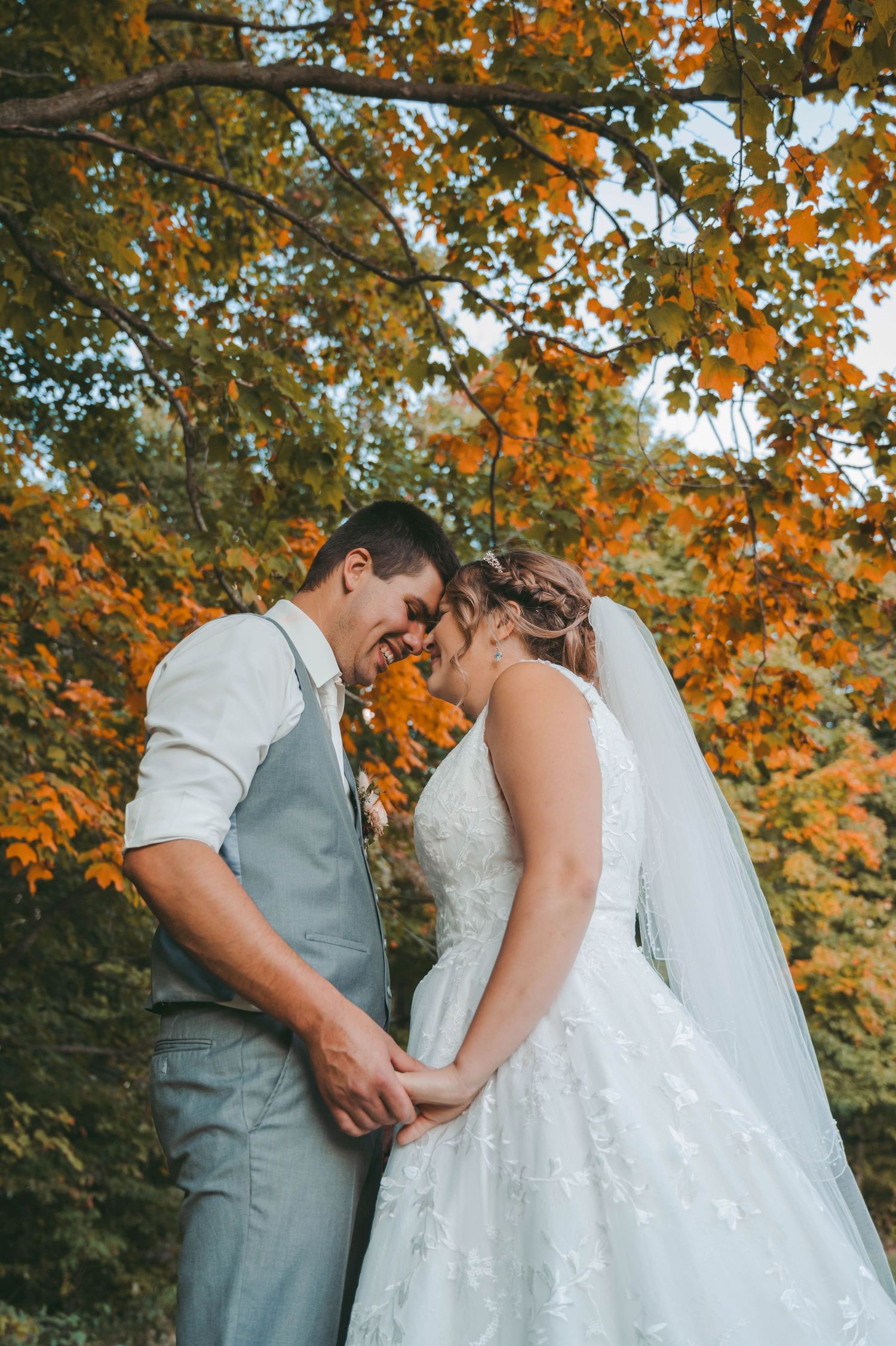 Intimate moment between couple in wedding attire under orange autumn tree branches.