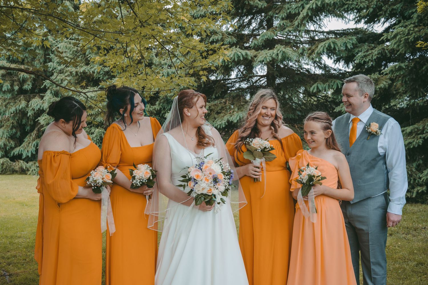 Wedding party with bridesmaids in matching orange dresses gather around bride holding bouquet outdoors.