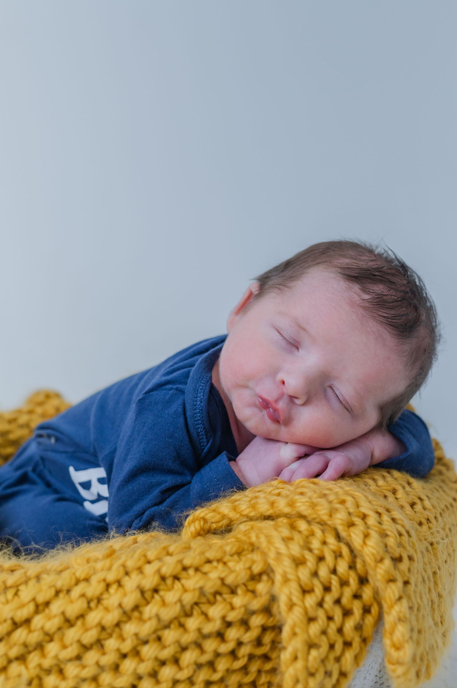 Sleeping newborn baby in blue onesie resting on yellow knit blanket against light gray background.
