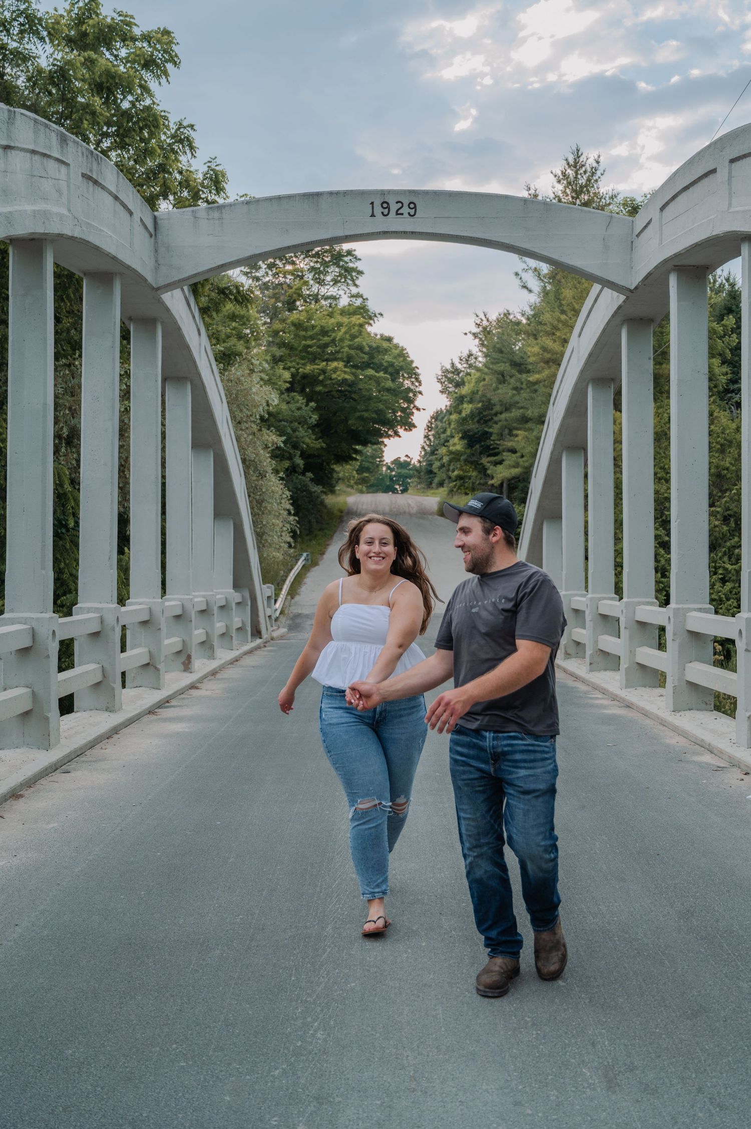 A couple walks together on a white arched bridge marked 1923 surrounded by green trees on a summer evening.