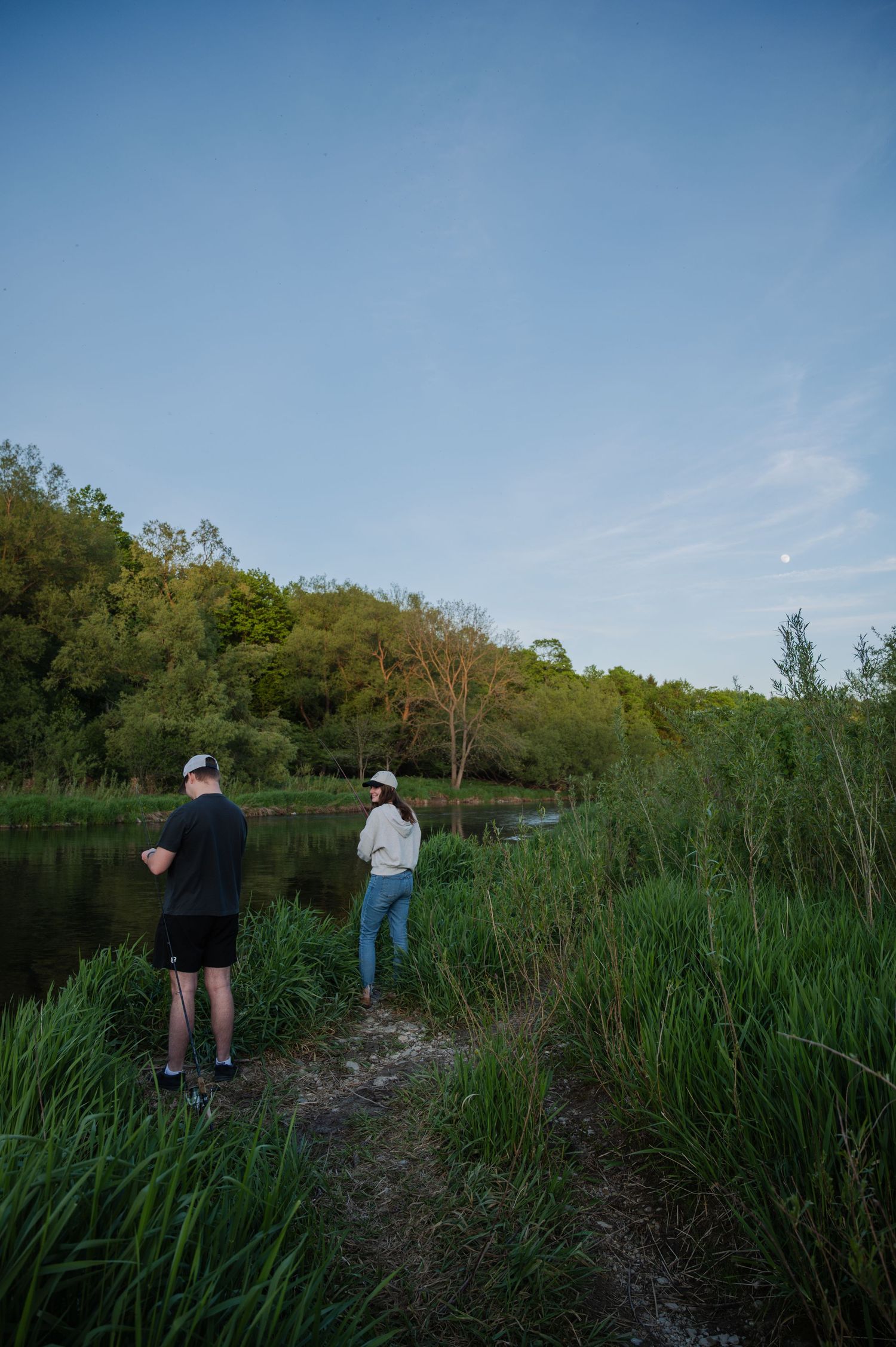 Two people fishing by a lush green riverbank at dusk with trees and a moonlit sky in the background.