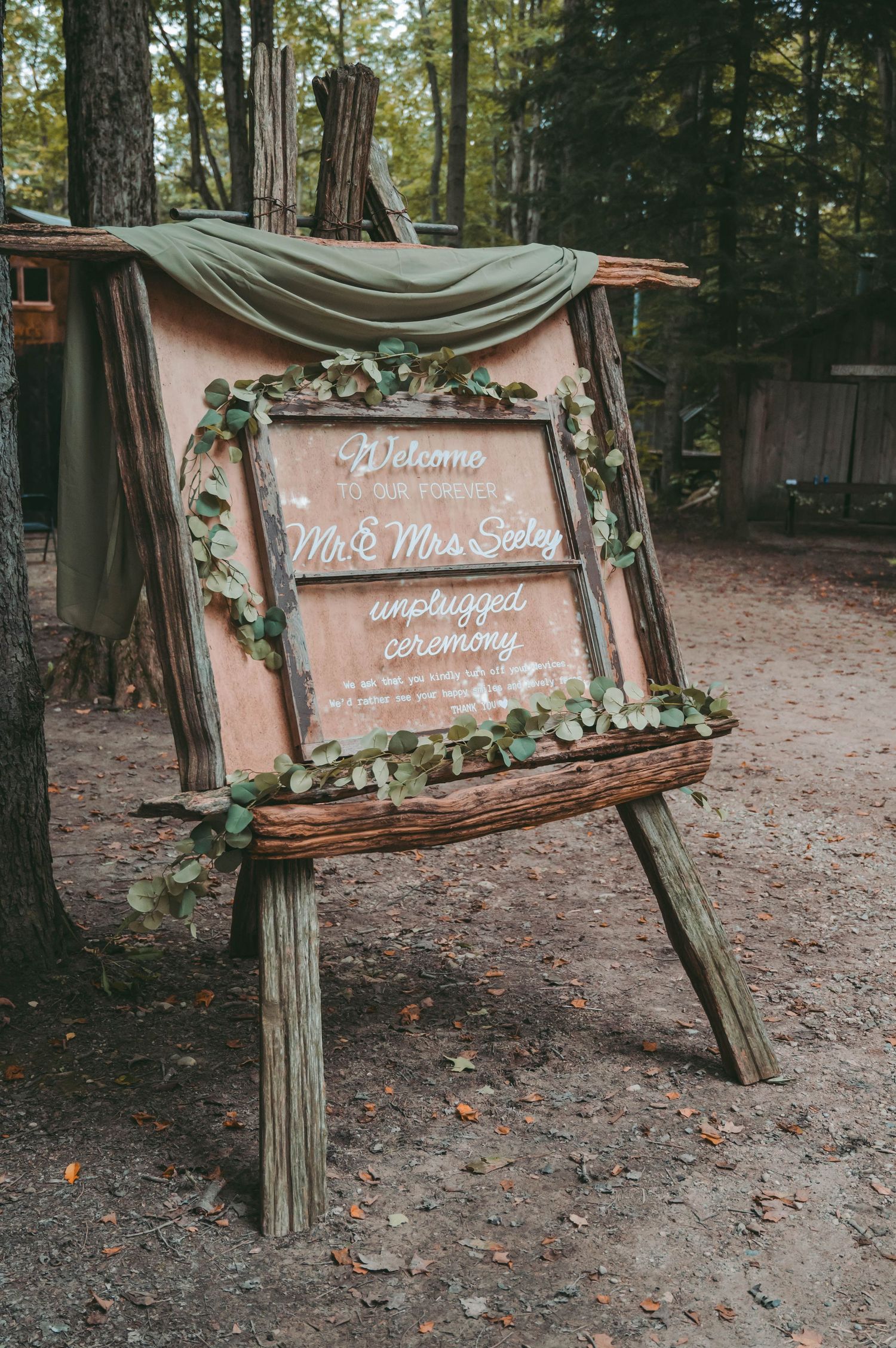A rustic wooden welcome sign decorated with greenery and fabric draping stands on an easel at an outdoor wedding ceremony.
