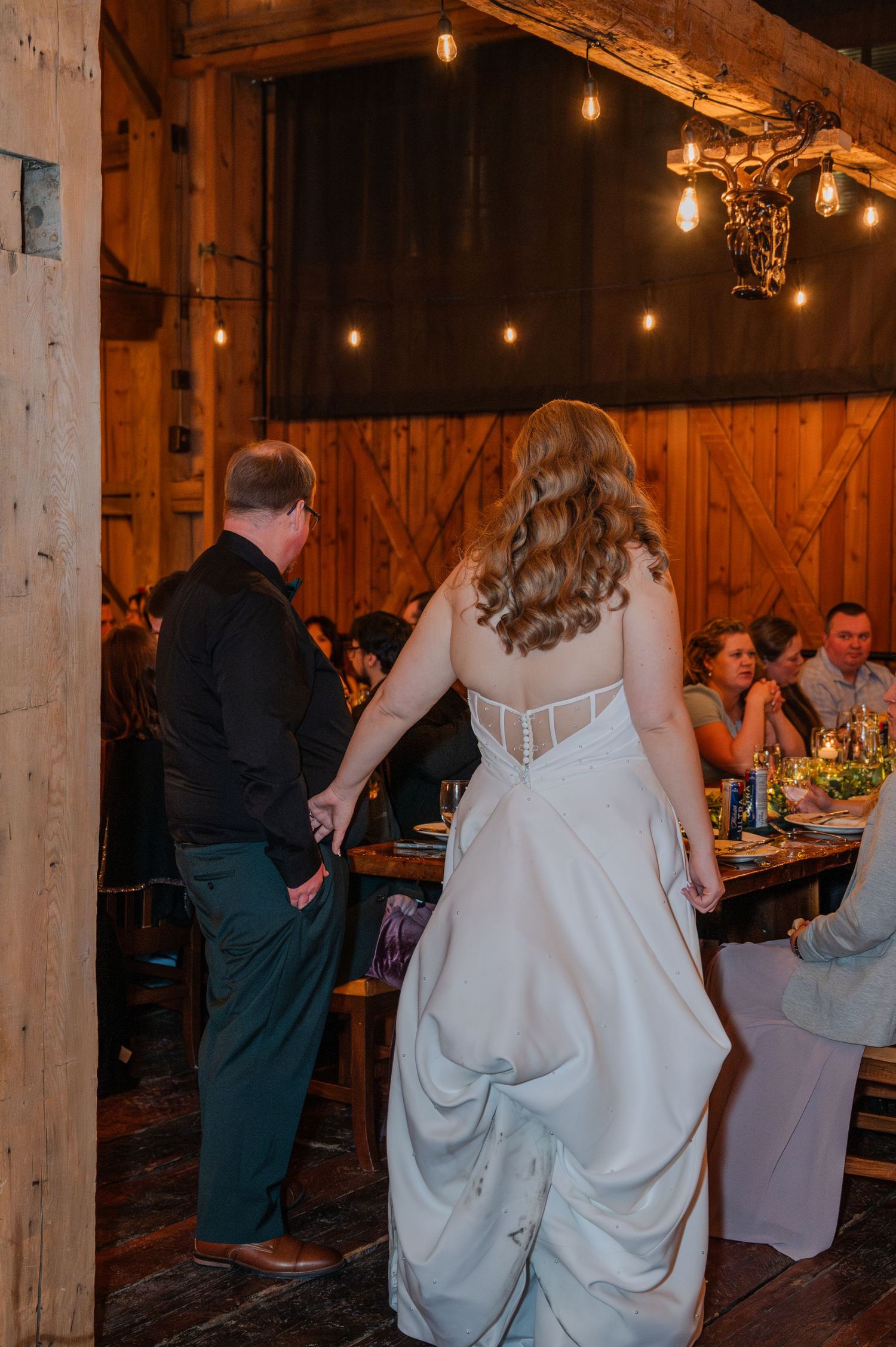 Bride in white gown and groom walking hand in hand inside a warmly lit rustic barn wedding reception.