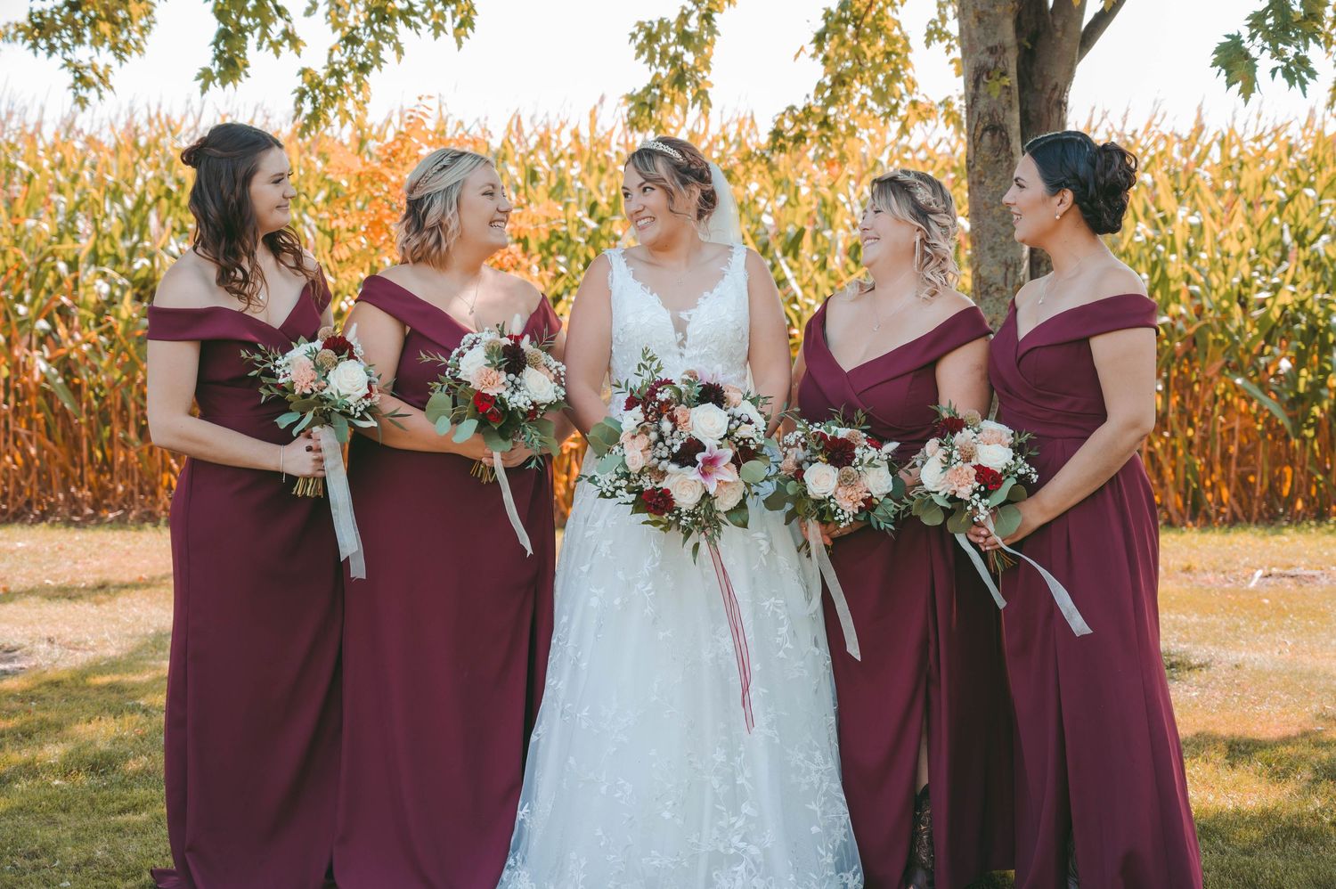 Bride in white gown poses with bridesmaids wearing burgundy dresses against autumn cornfield backdrop.