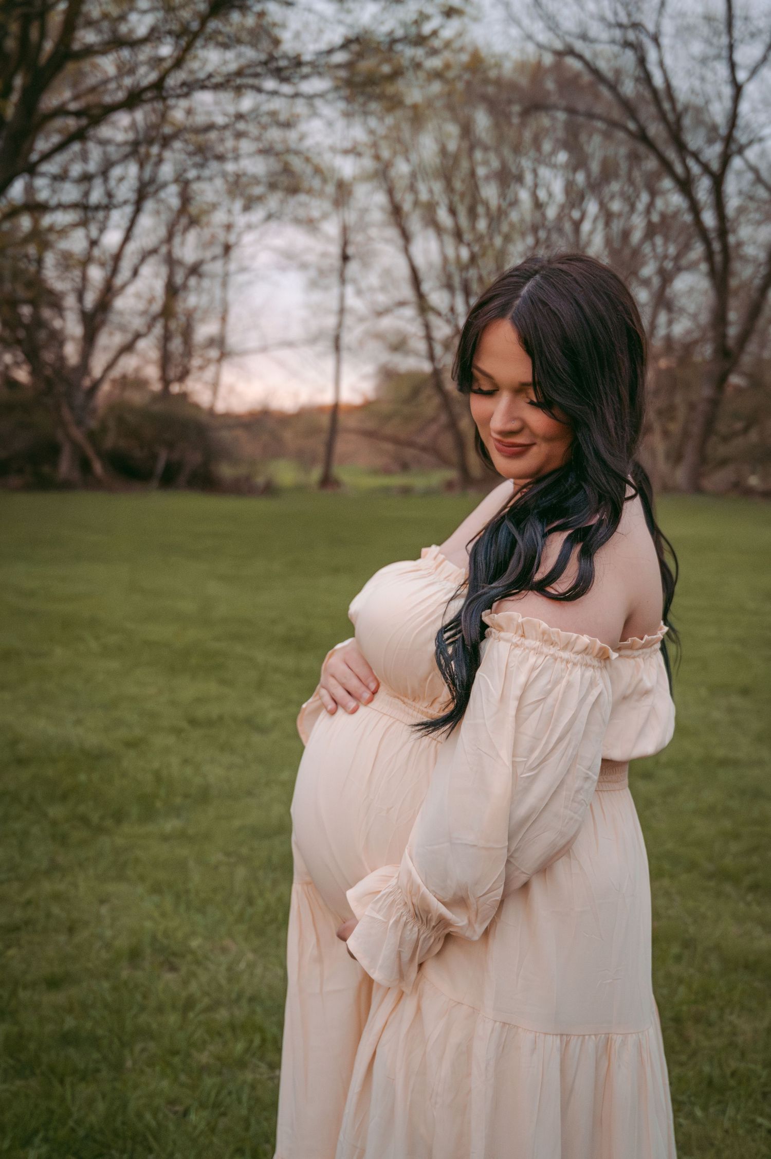 Expectant mother in off-shoulder cream dress smiles while touching belly during golden hour.