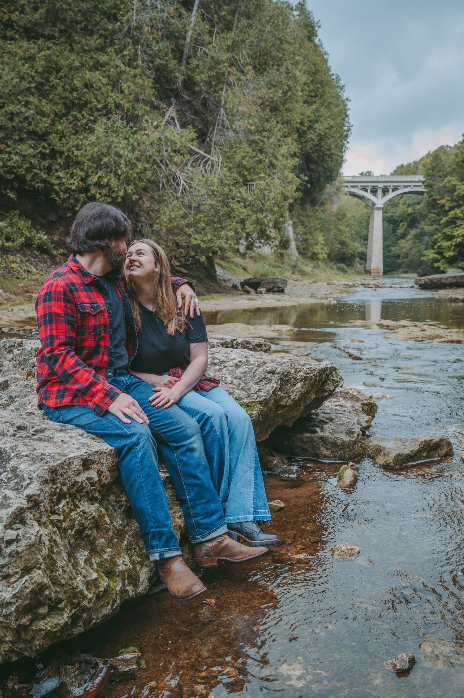 Couple sharing intimate moment while sitting on rocks by river with the Elora Gorge bridge in background.
