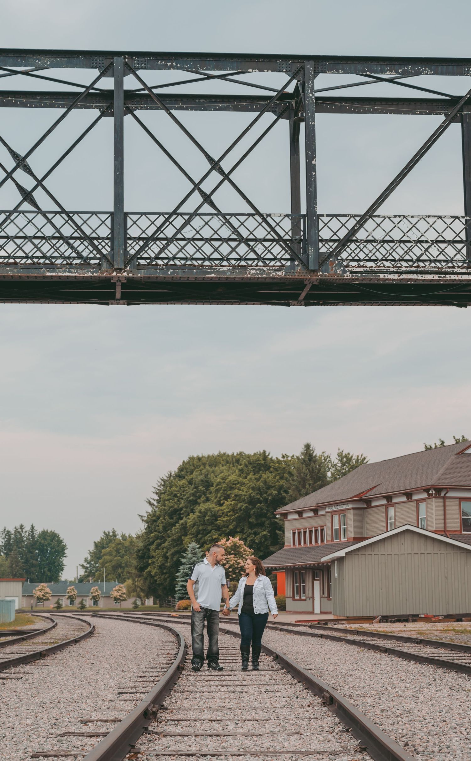 A couple walking hand in hand along railroad tracks beneath a large metal bridge structure.