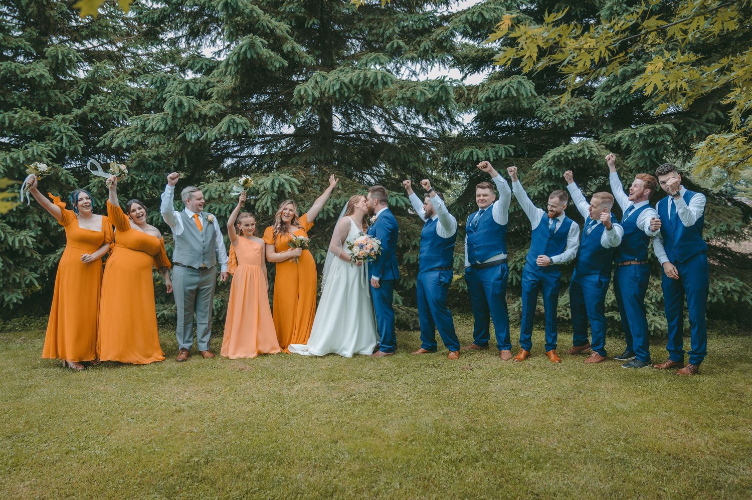 Wedding party in orange and blue attire poses cheerfully on lawn with evergreen backdrop.