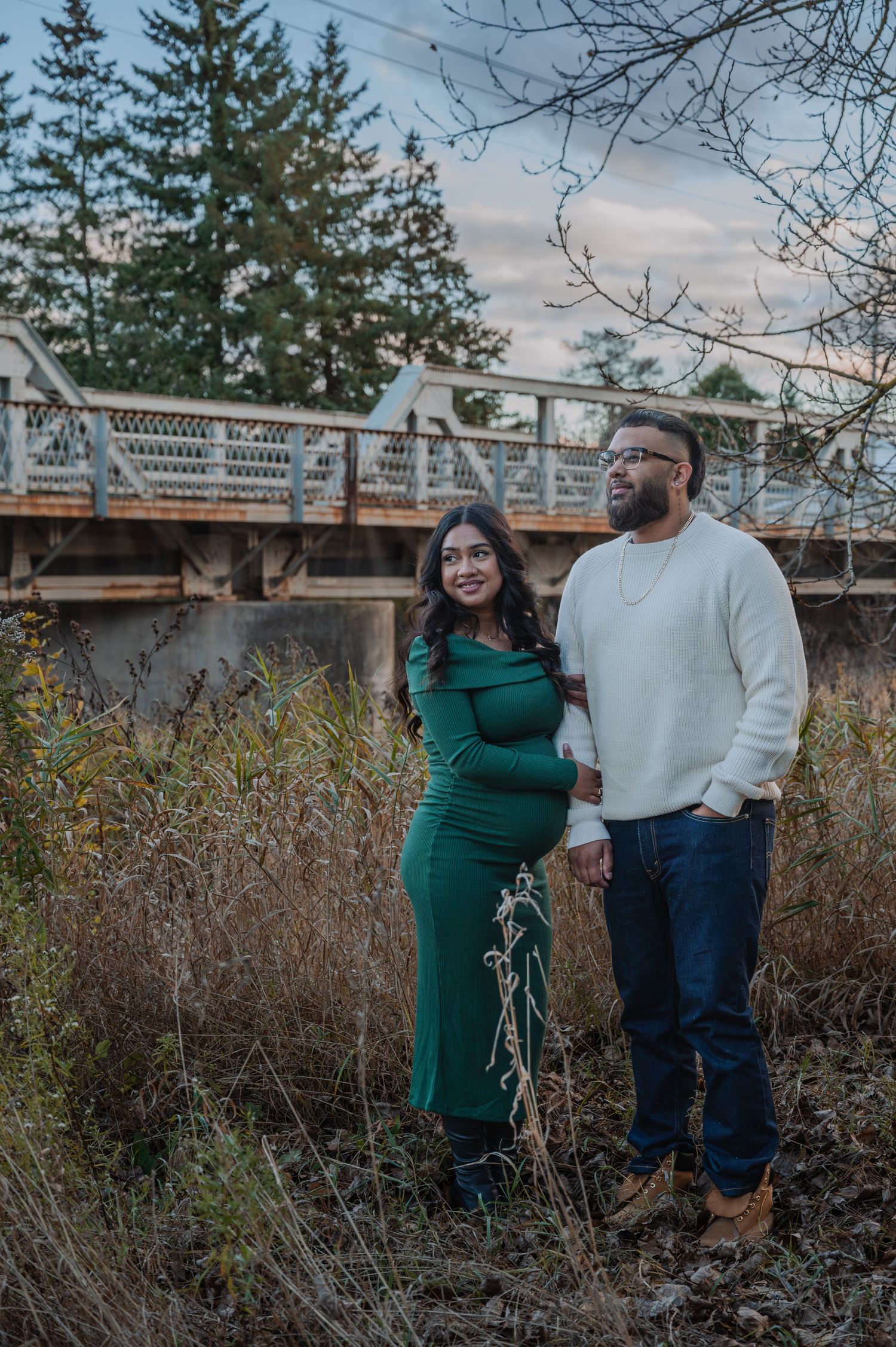 A couple stands together in a rustic outdoor setting with tall grass and evergreen trees in the background.