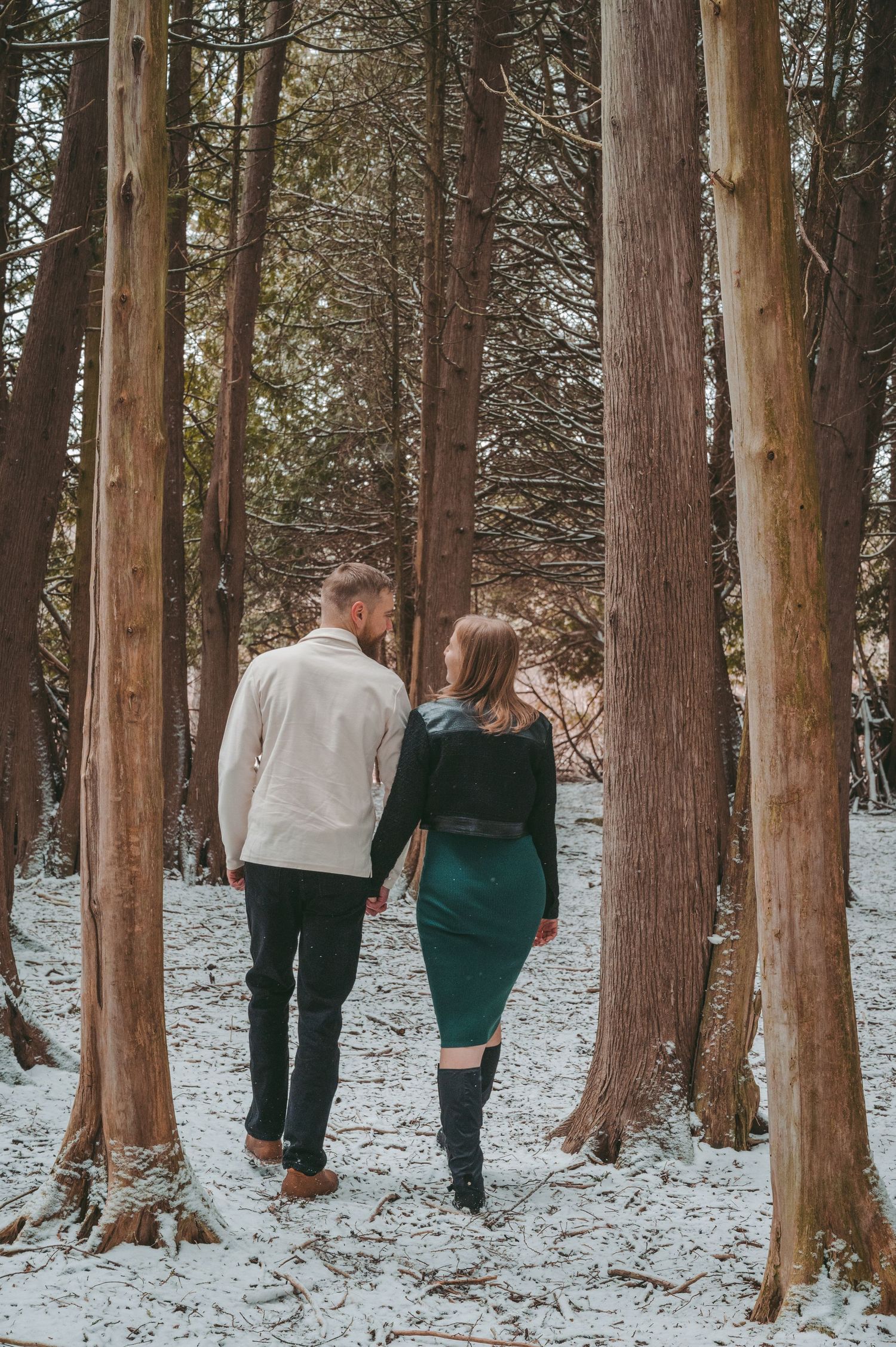 A couple walks through a snowy forest of tall cedar trees on a winter day.