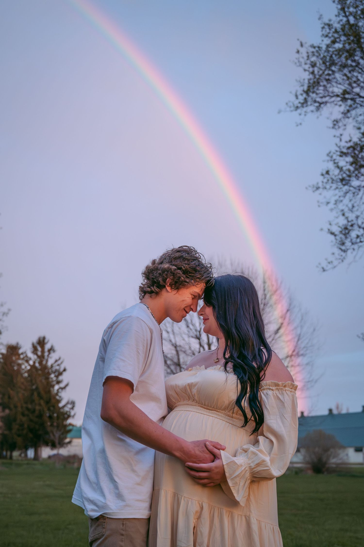 Couple embraces under a vibrant rainbow during outdoor maternity photoshoot.