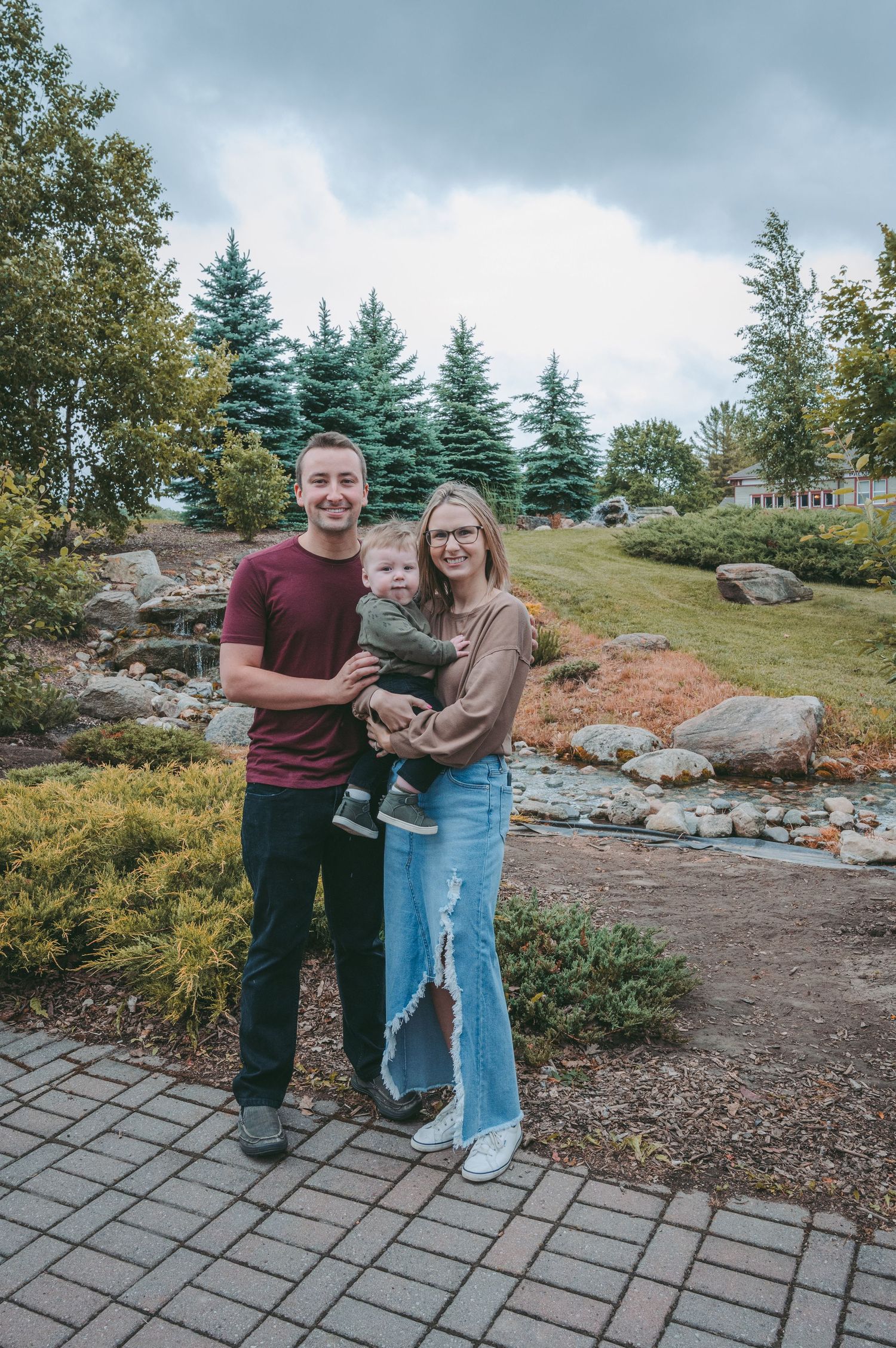 Couple standing together on landscaped path with evergreen trees in background.