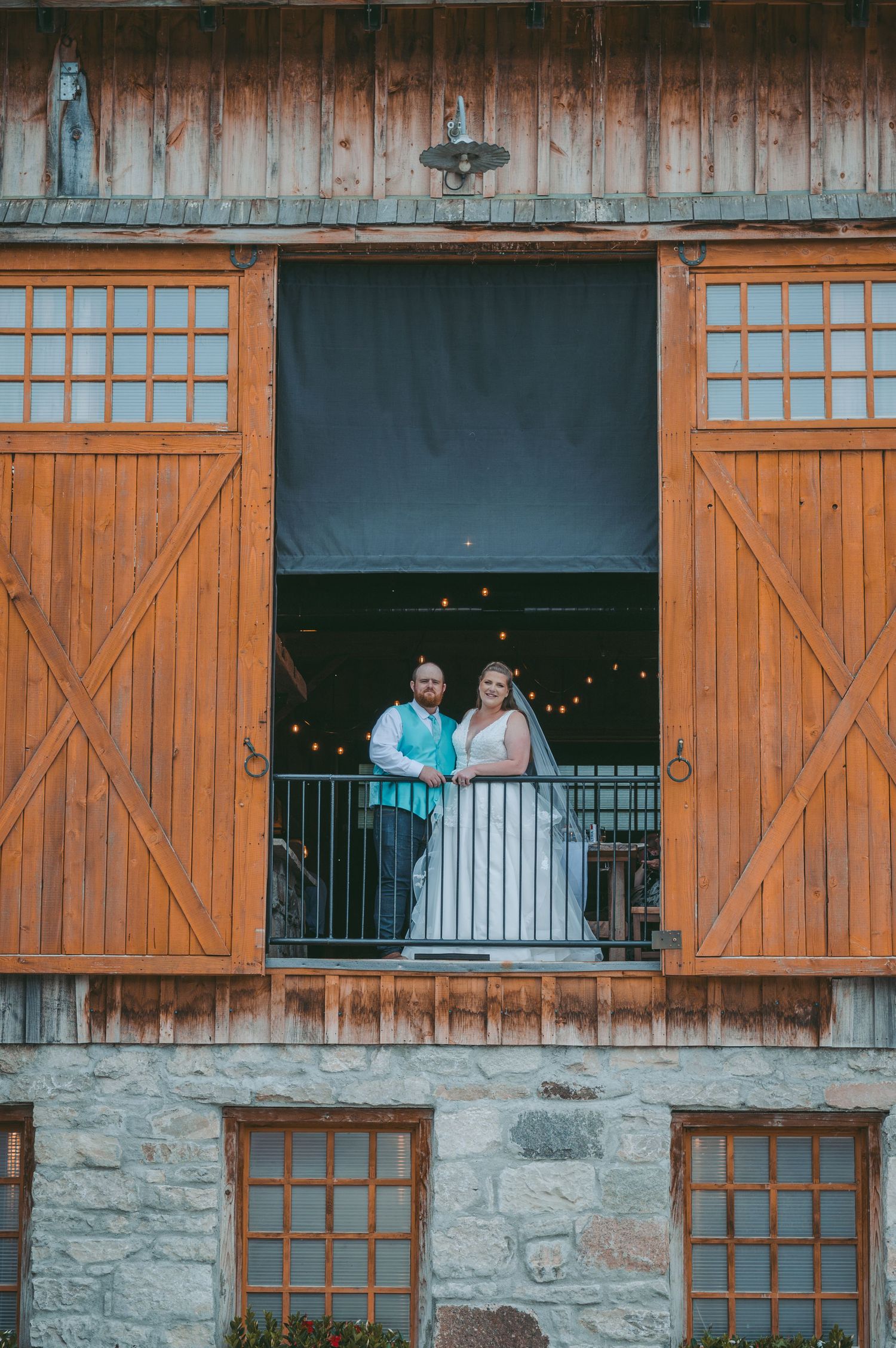 A couple stands in formal attire on a balcony of a rustic barn with wooden doors and string lights.