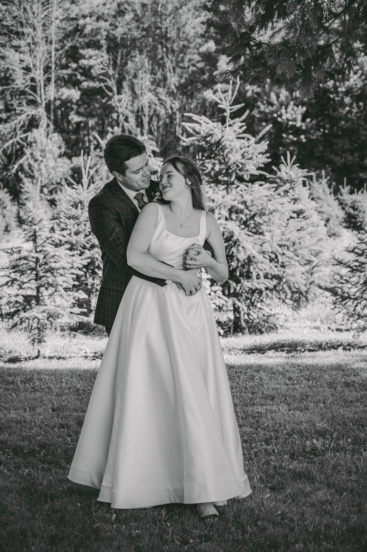 Black and white portrait of newlyweds embracing in front of evergreen trees.