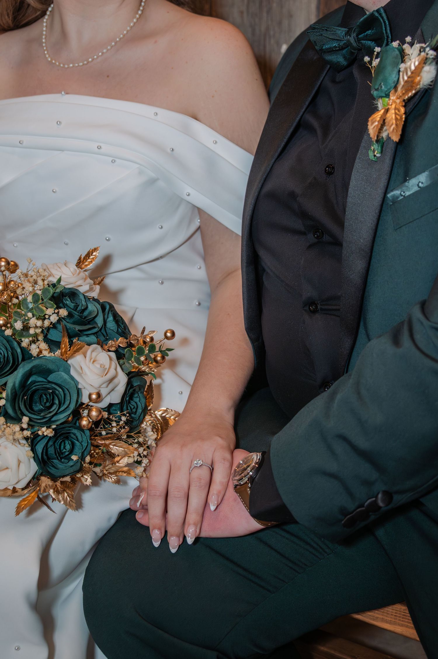 Close-up of wedding couple's hands with teal and gold floral details on white dress against dark suit.