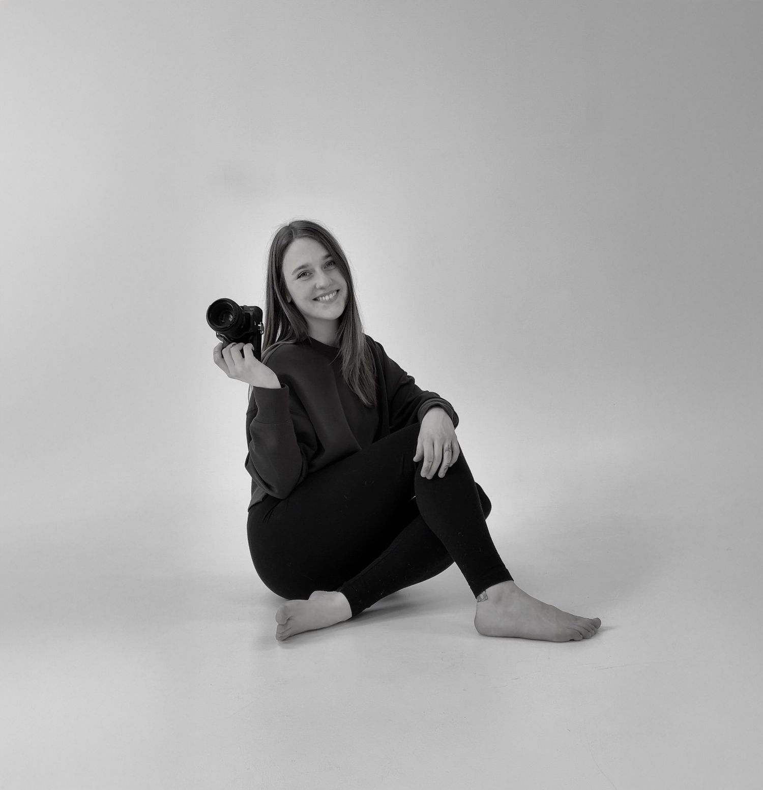 Black and white photo of a smiling young woman sitting on floor holding a camera, wearing dark casual outfit, barefoot.