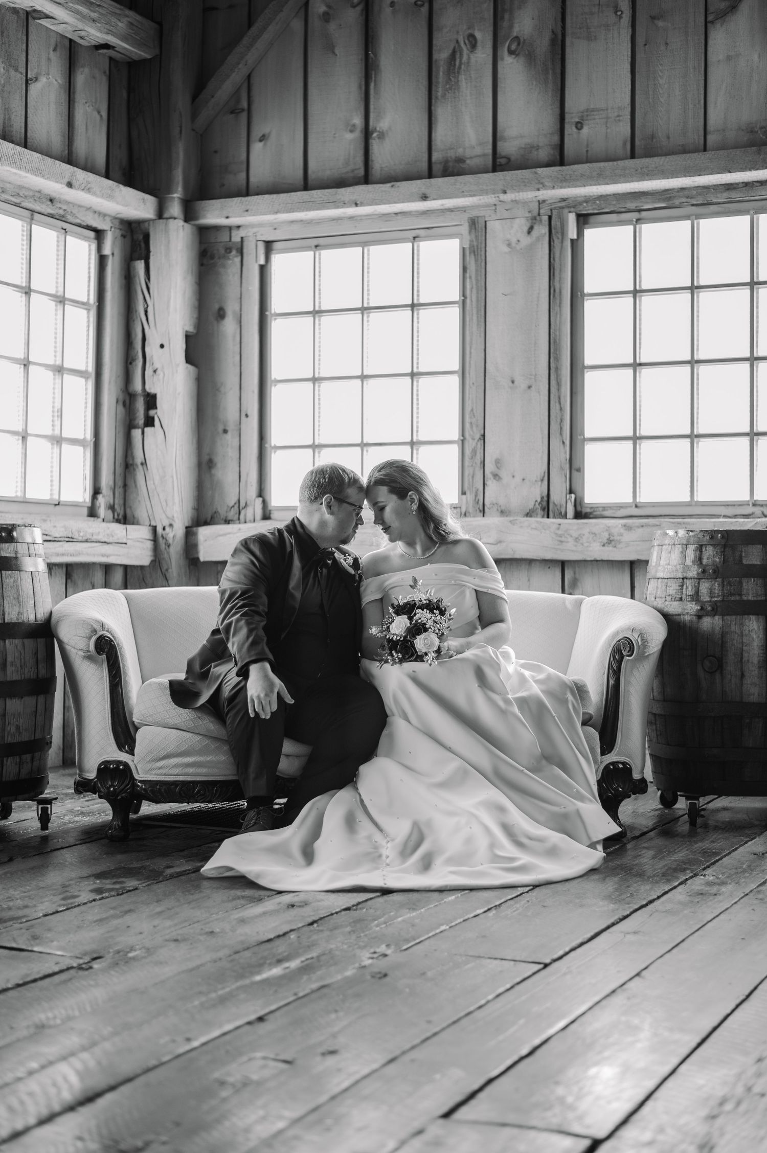 Black and white photo of bride and groom sharing an intimate moment on a vintage sofa in a rustic barn venue.