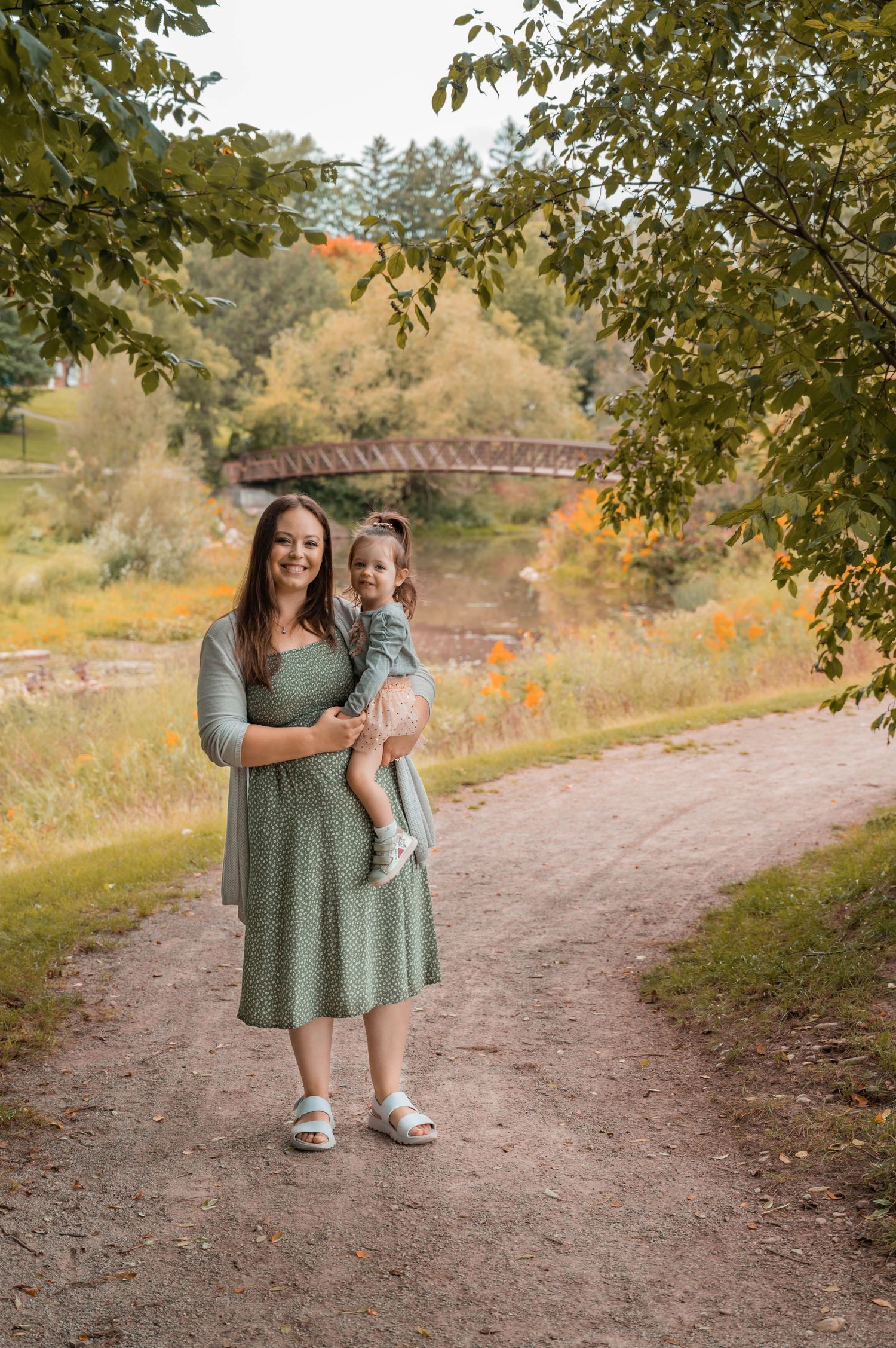 Two people share a tender moment on a dirt path with a rustic bridge and autumn scenery in the background.