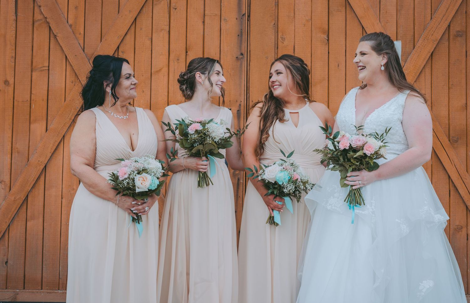 Bridesmaids in blush dresses hold bouquets while standing against a wooden barn door.