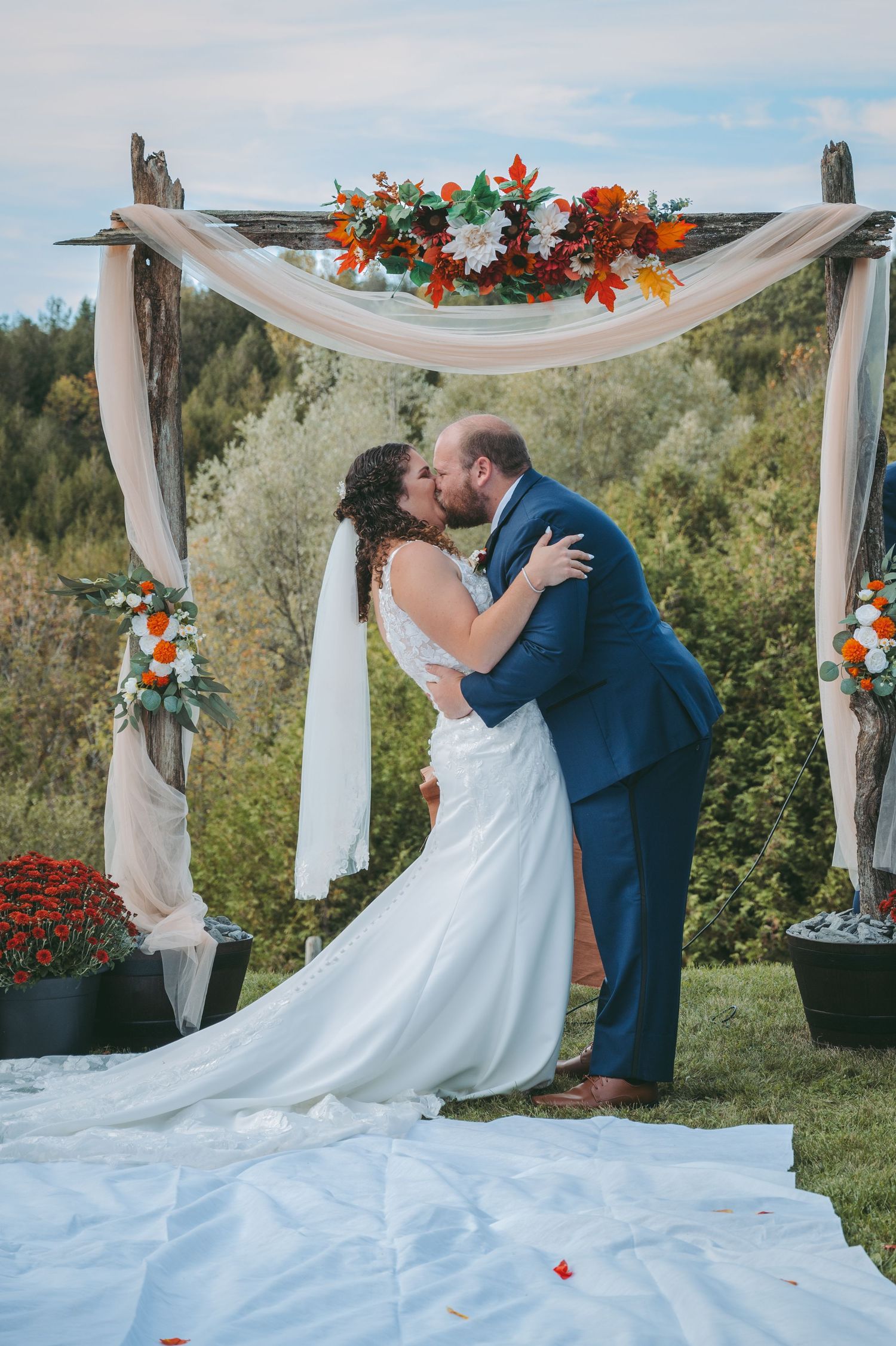 Couple shares a romantic kiss under a floral arch with white draping and orange flowers during outdoor wedding ceremony.