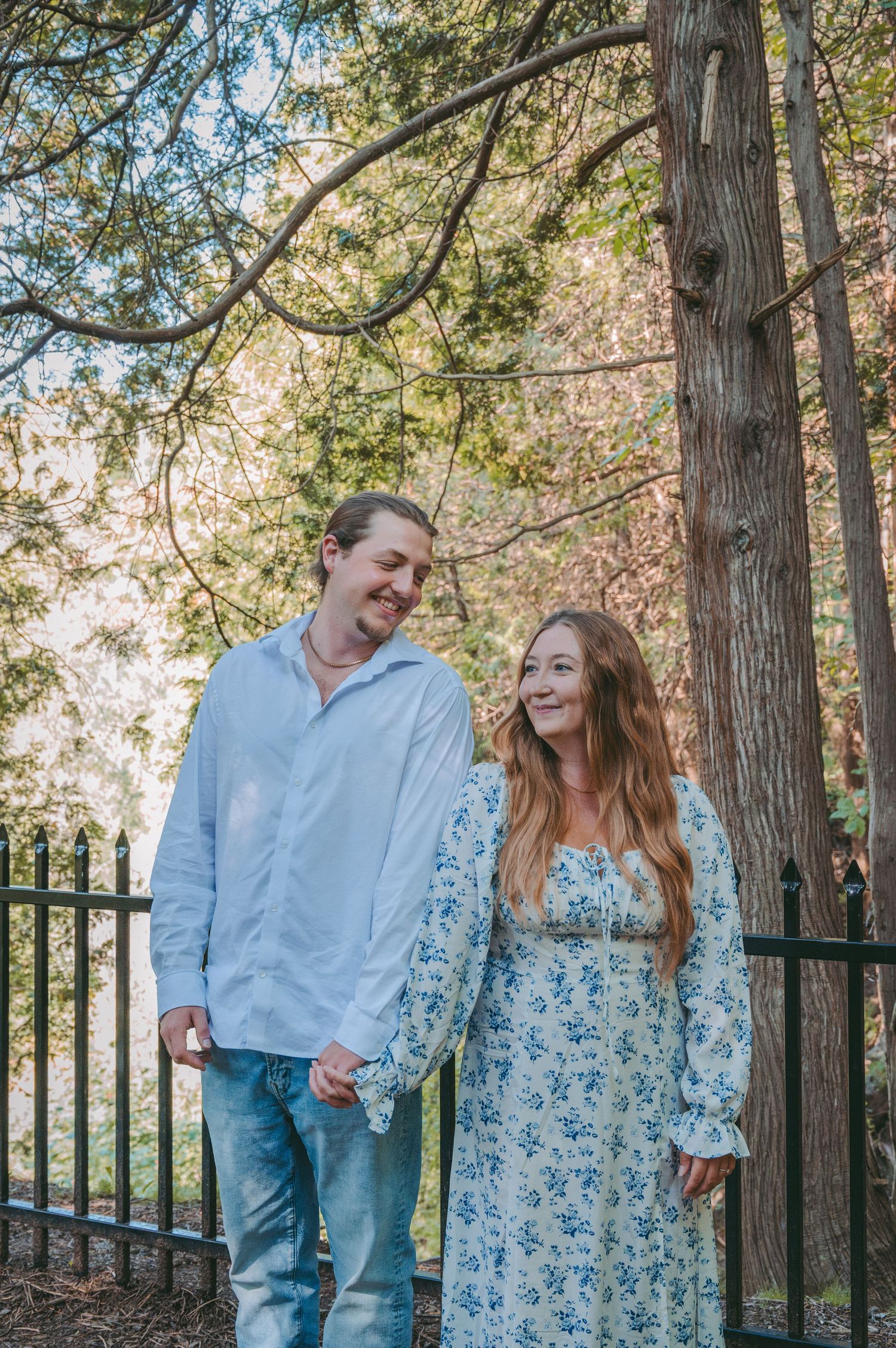 A couple laughs together while standing among tall trees and an iron fence.