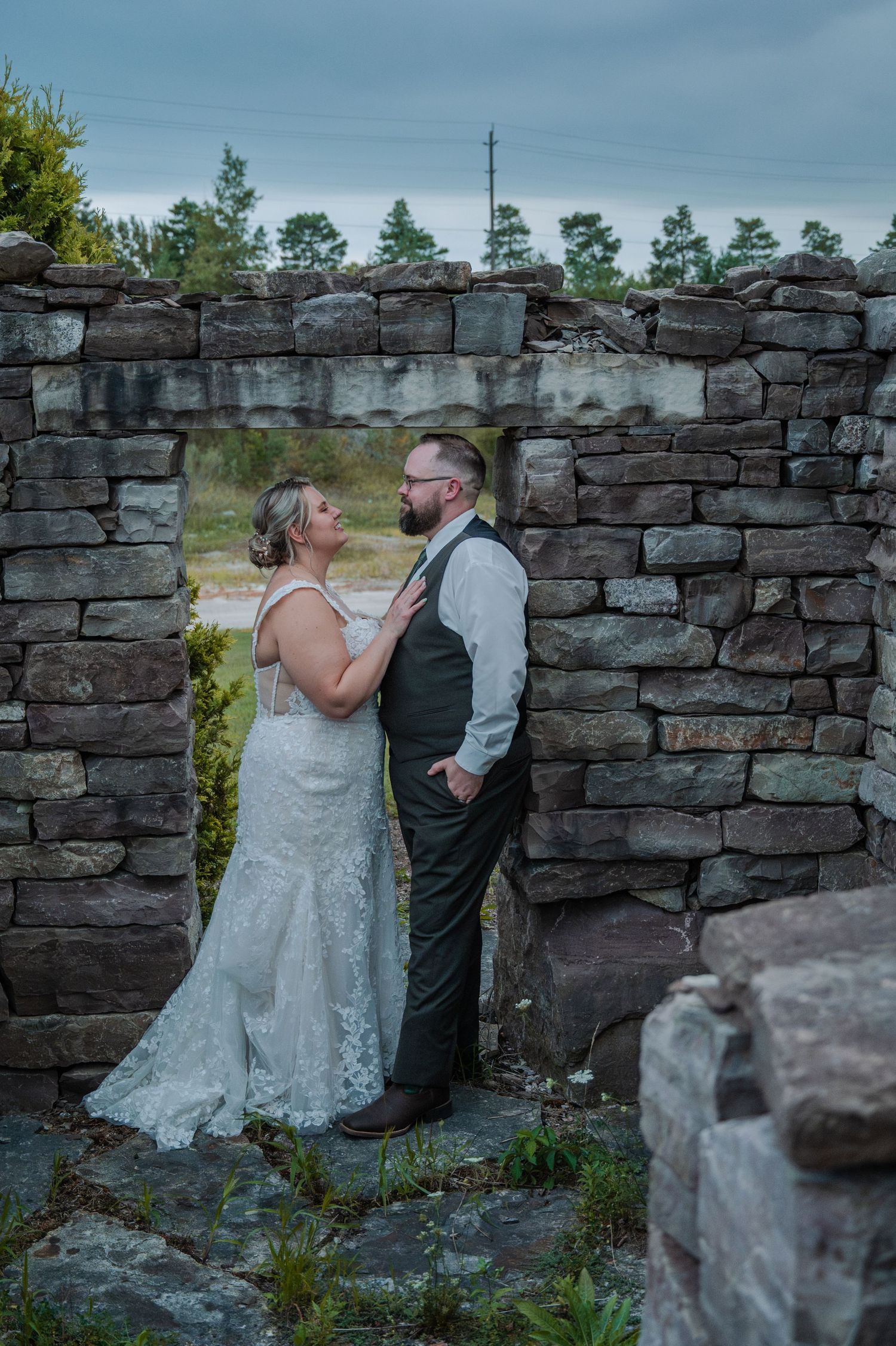 Bride and groom share a romantic moment in a rustic stone archway, bride in lace gown, groom in gray vest.