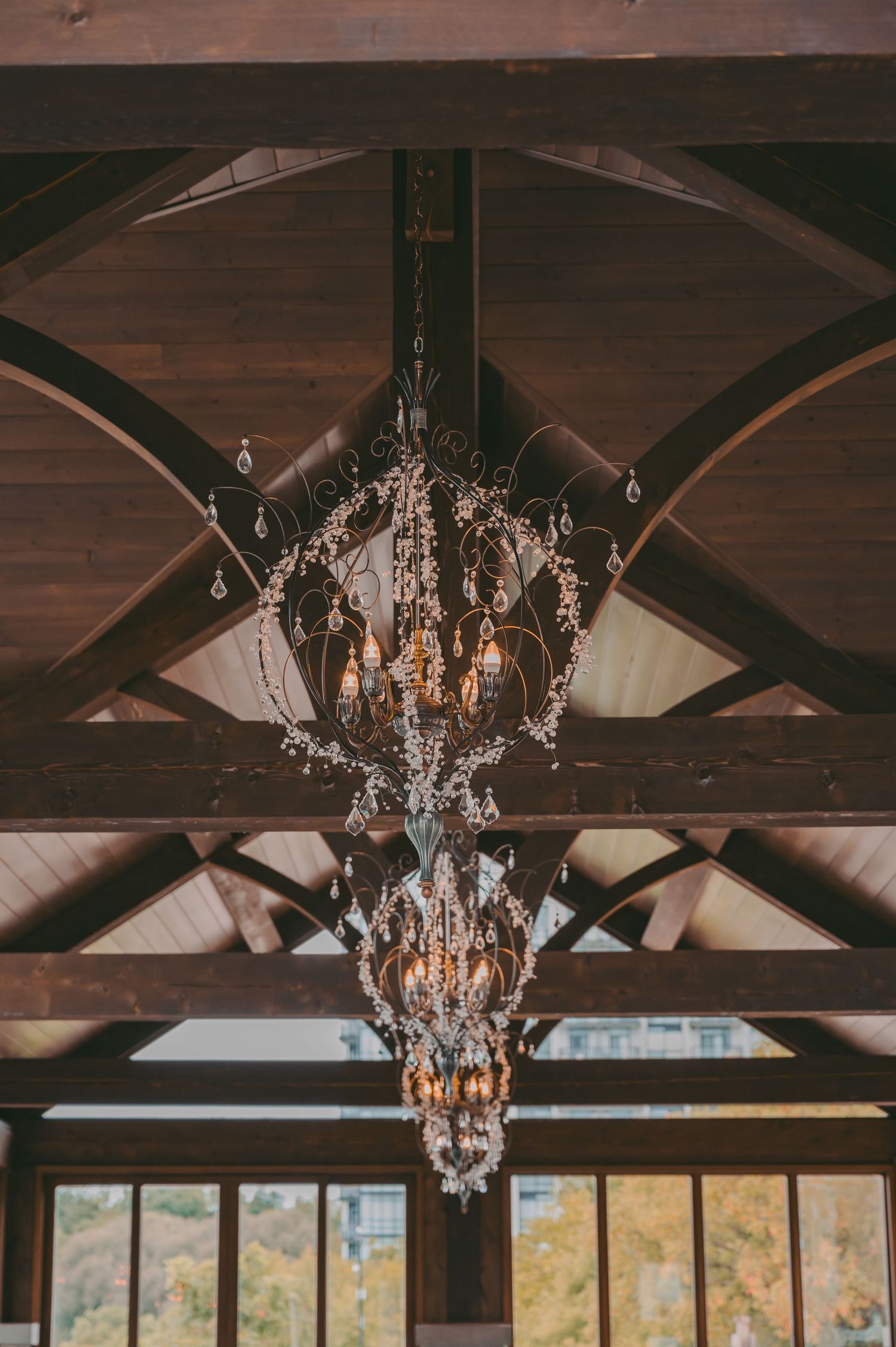 Elegant crystal chandeliers hang from dark wooden beams in the Cambridge Mill with autumn views through windows.