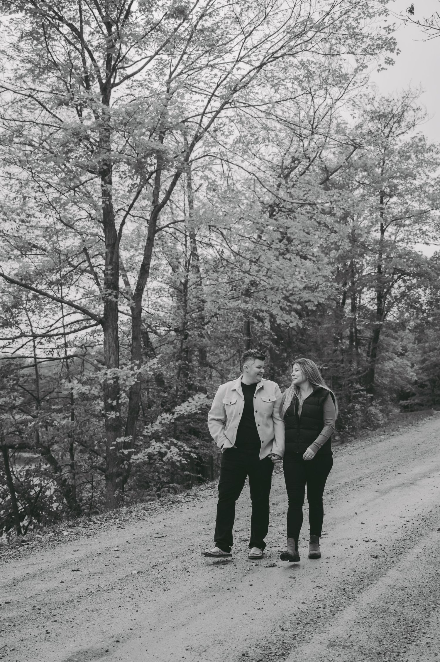 A couple walks together on a dirt path through autumn woods in black and white.