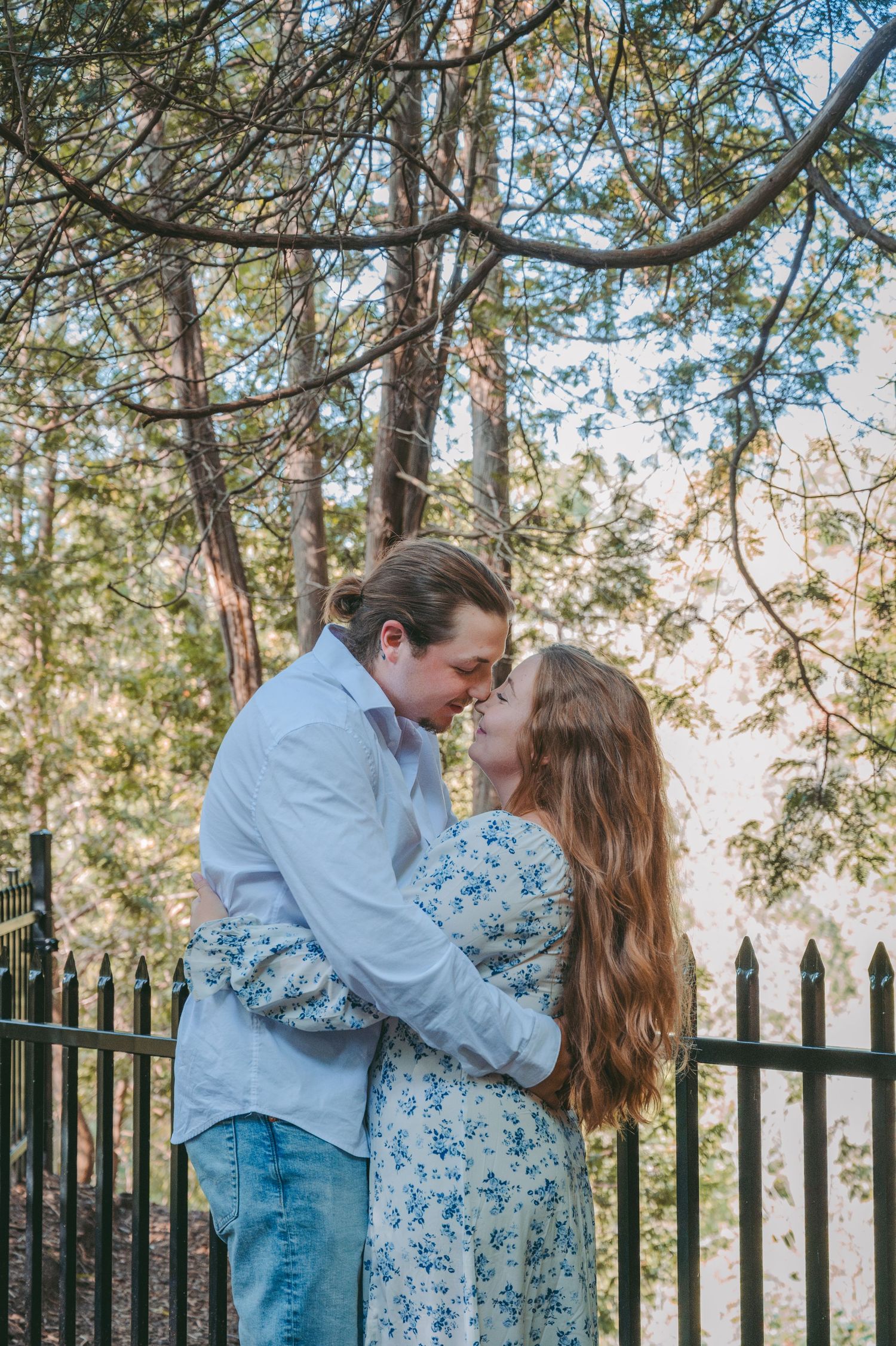 A couple embraces tenderly next to a black iron fence in a wooded setting.