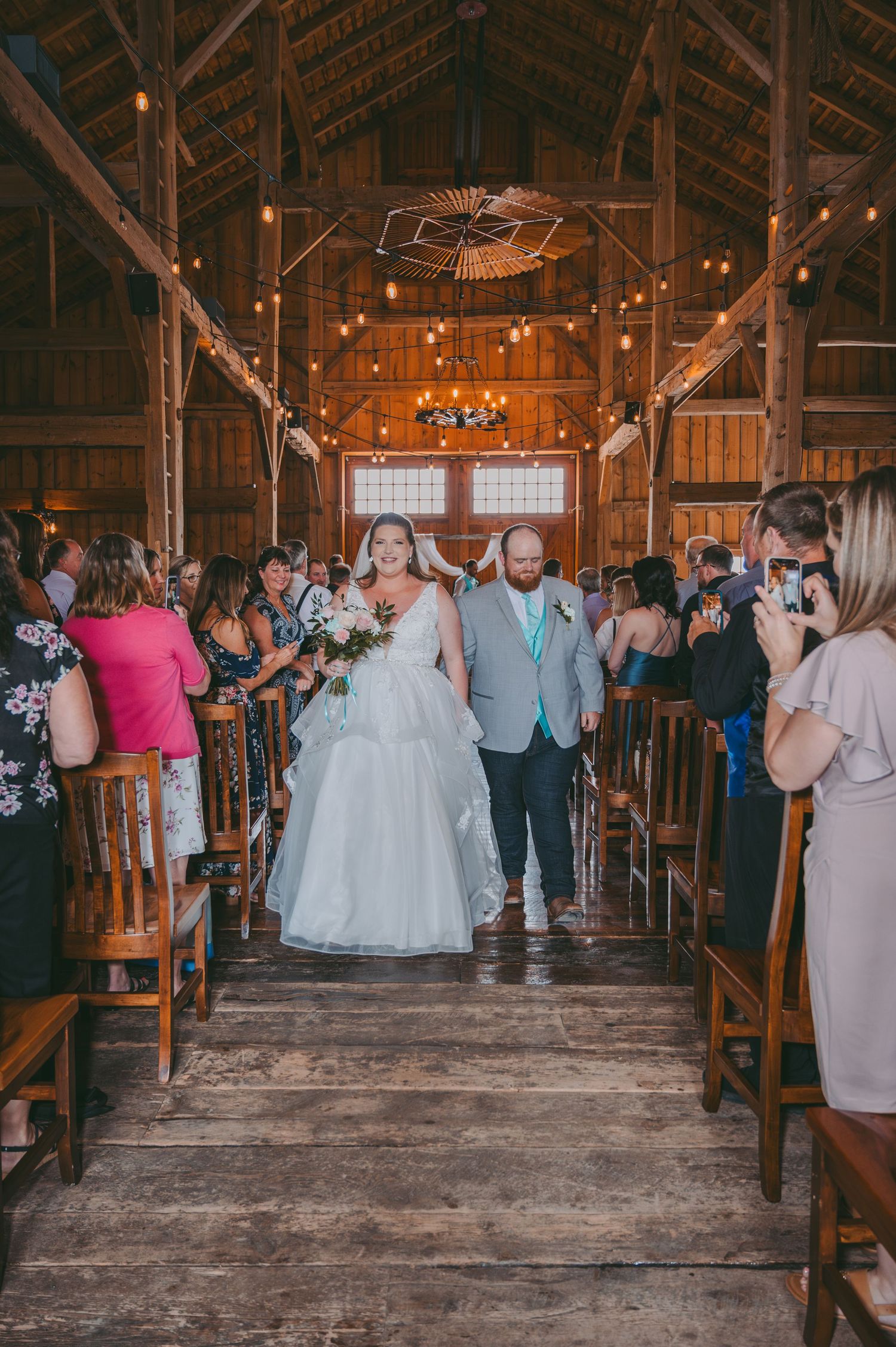 Newlyweds exit their barn wedding ceremony while guests line the aisle with cameras.