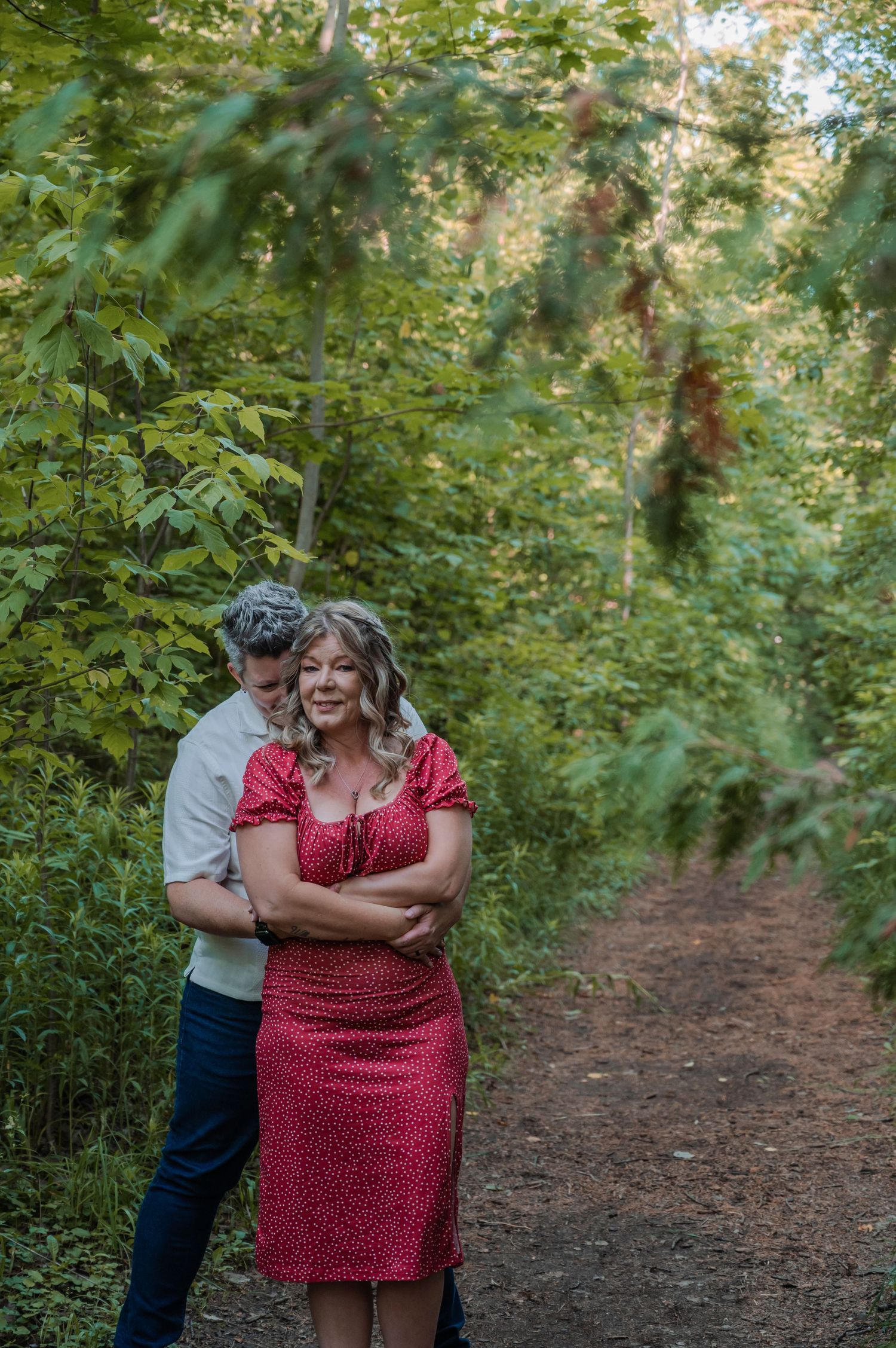 Couple shares an intimate embrace on a wooded path surrounded by lush green trees and foliage.