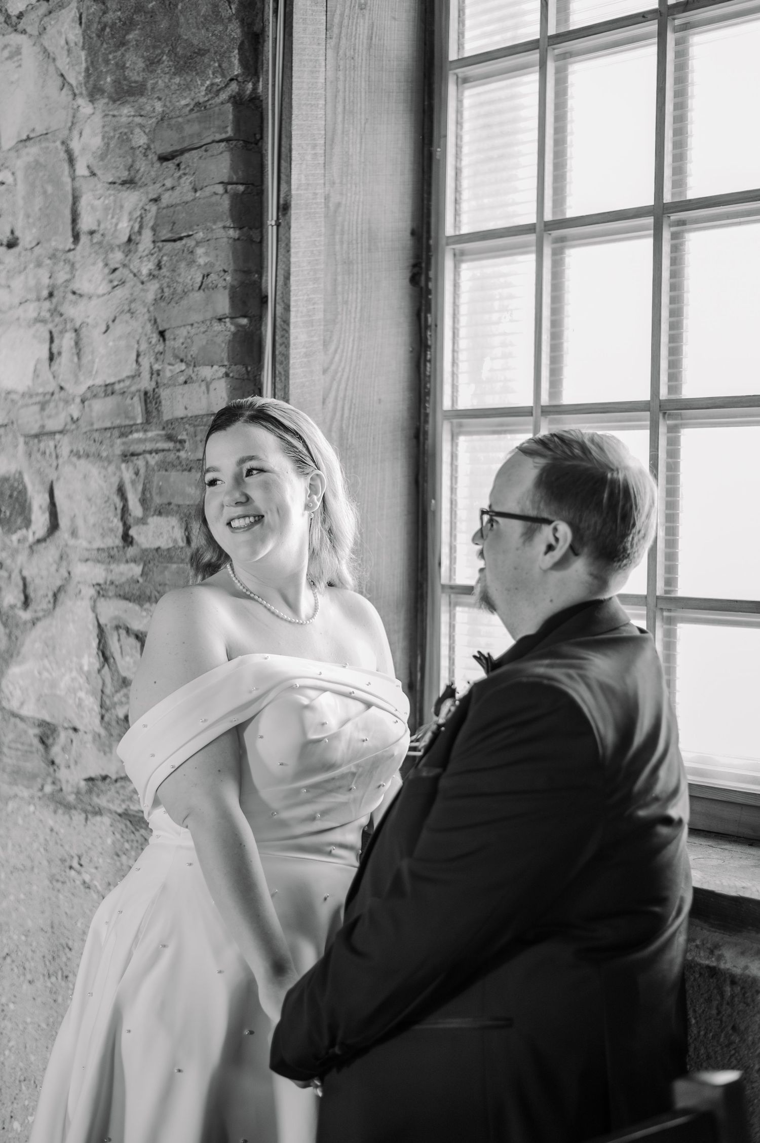 Black and white wedding photo of smiling bride in strapless gown and groom in suit seated by large window.