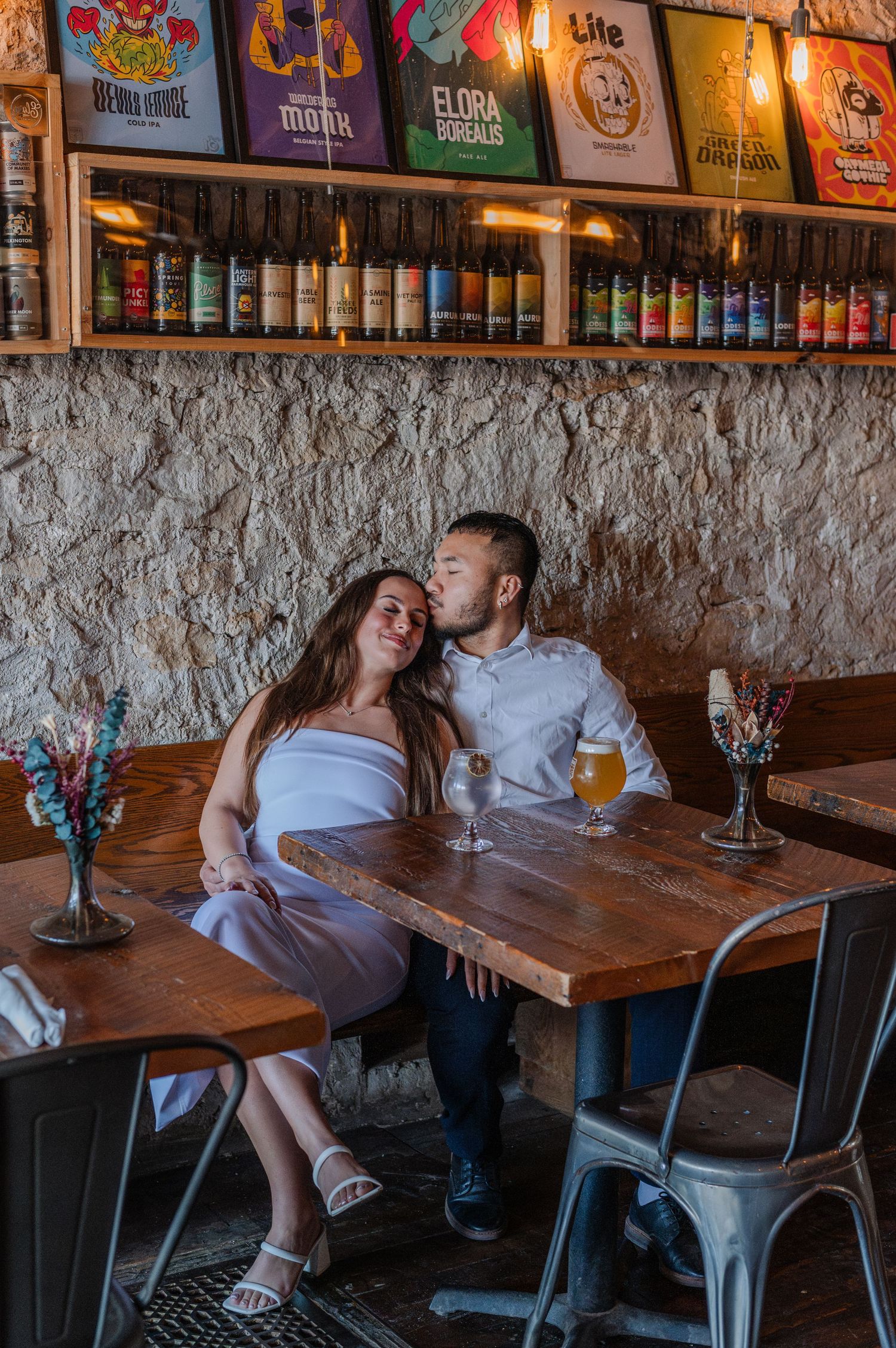 Couple sharing a romantic kiss at a rustic brewery bar table with craft beer bottles displayed on shelves behind them.