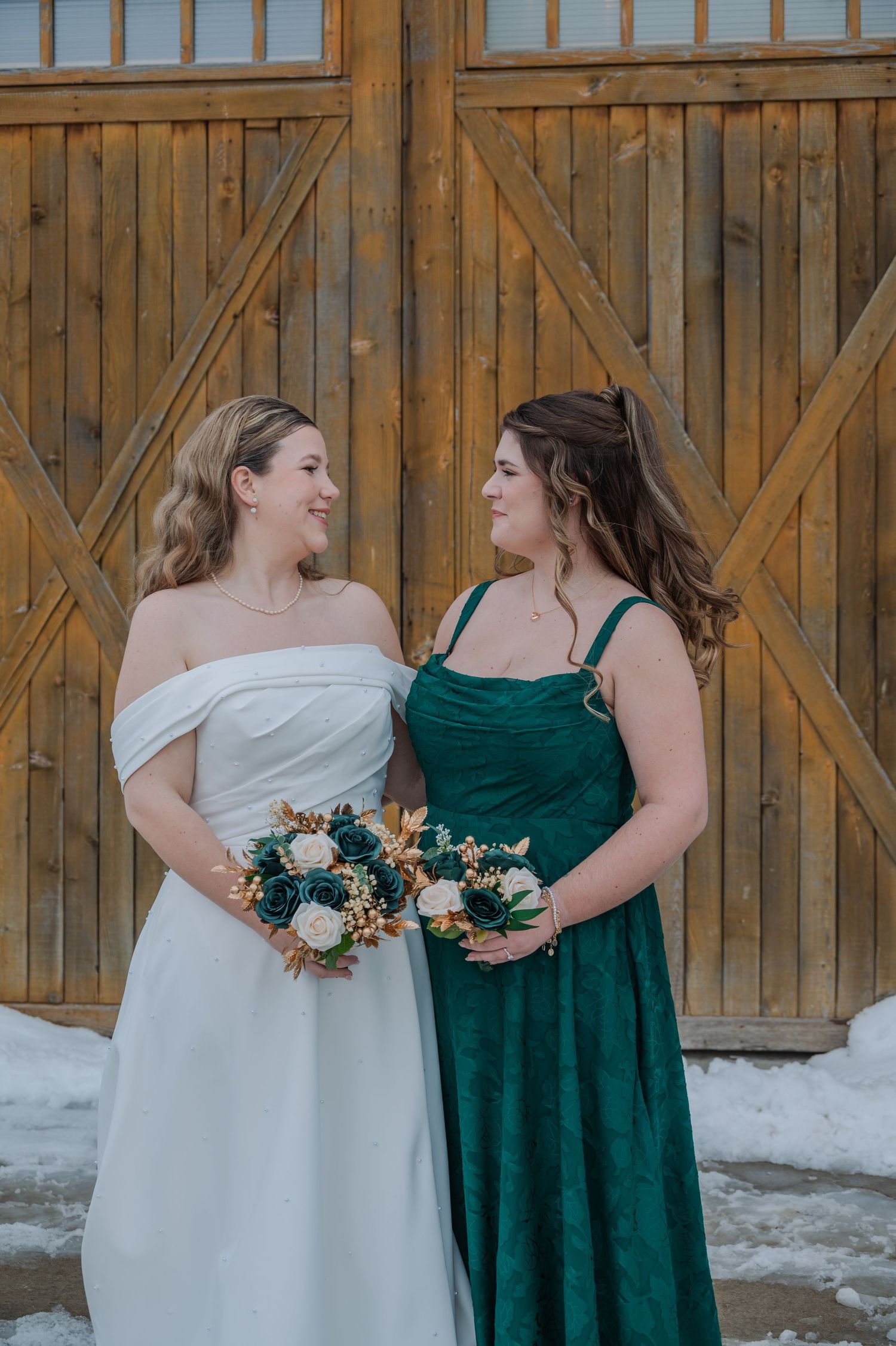 Bride in white off-shoulder gown and bridesmaid in emerald green dress holding bouquets before rustic barn doors in winter.