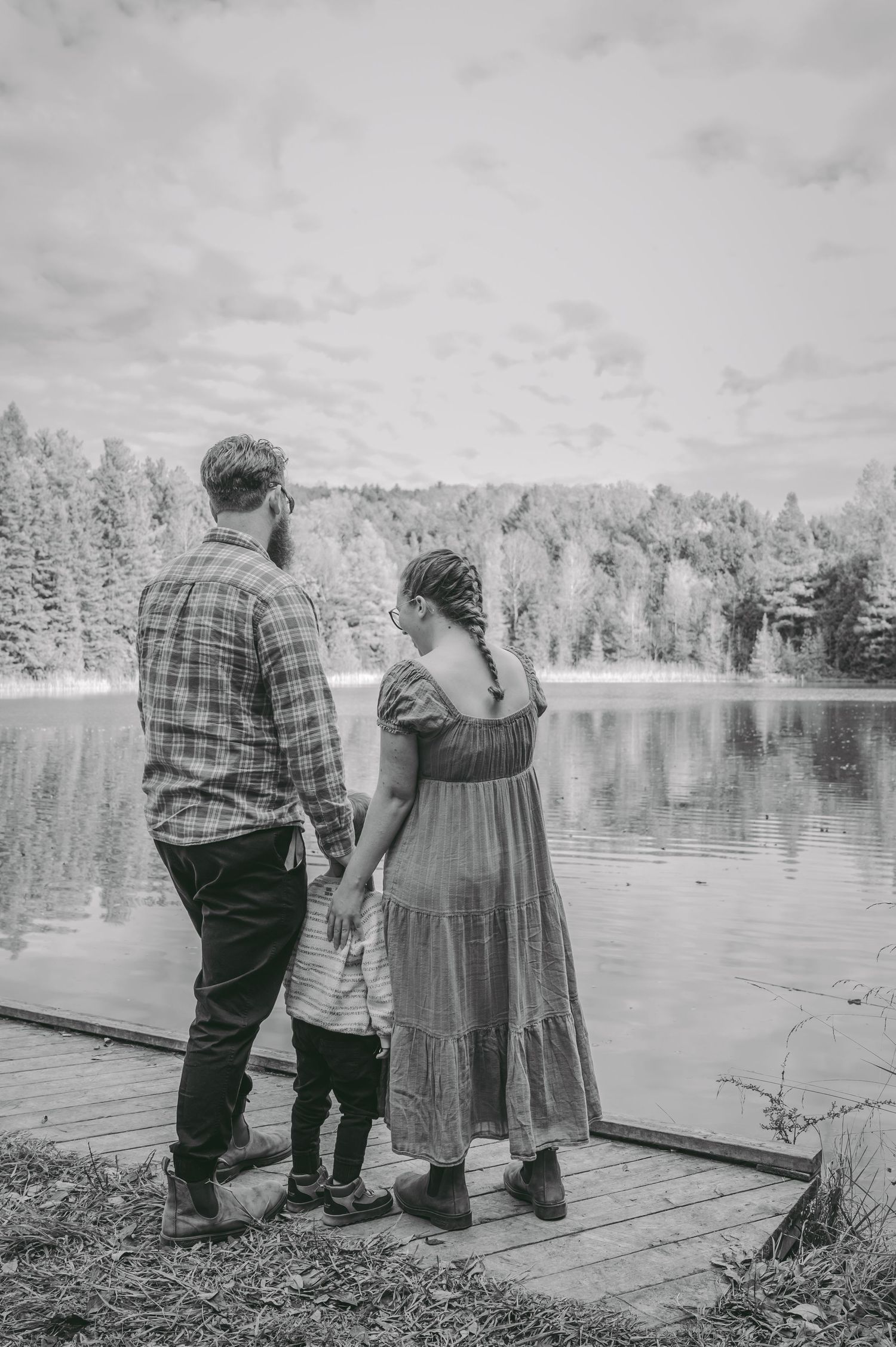 Family holds hands while standing on dock overlooking serene lake and forest in black and white.