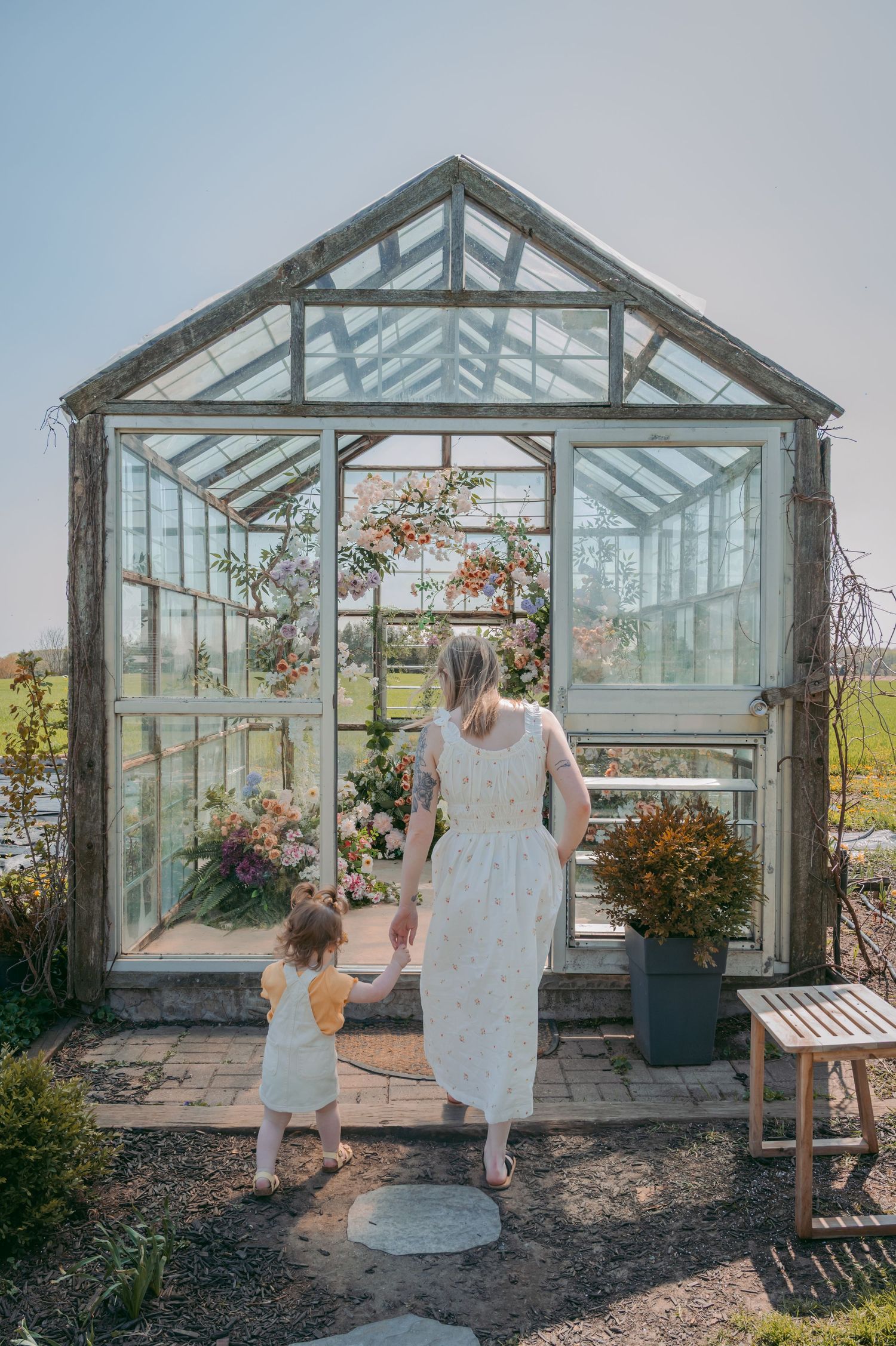 Two figures in white dresses stand before a glass greenhouse surrounded by natural gardens and sunlight.