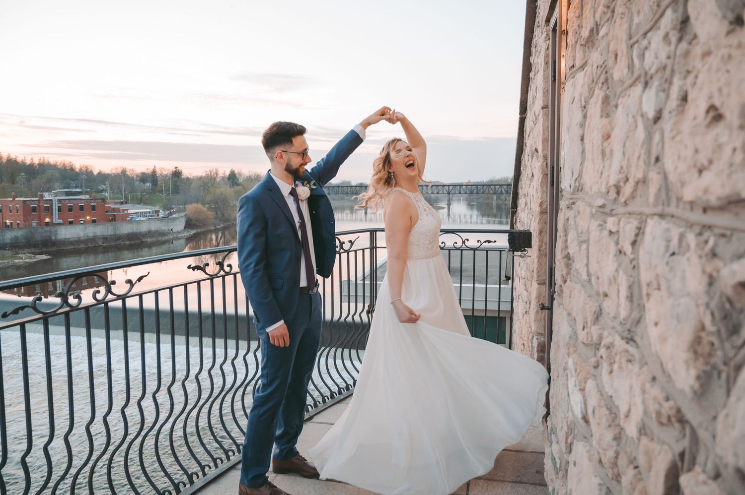Joyful wedding couple dancing on stone balcony with waterfront view.