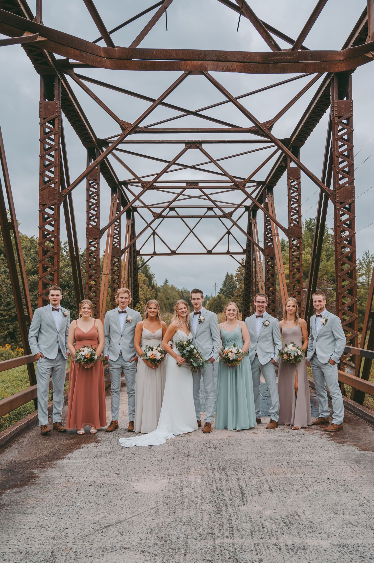 Wedding party in gray suits and pastel dresses posed on rustic metal bridge.