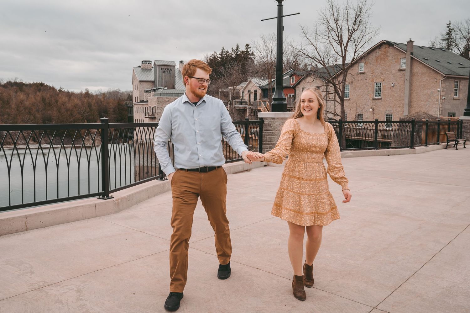 Two people stroll hand in hand along a waterfront promenade near an old flour mill building.