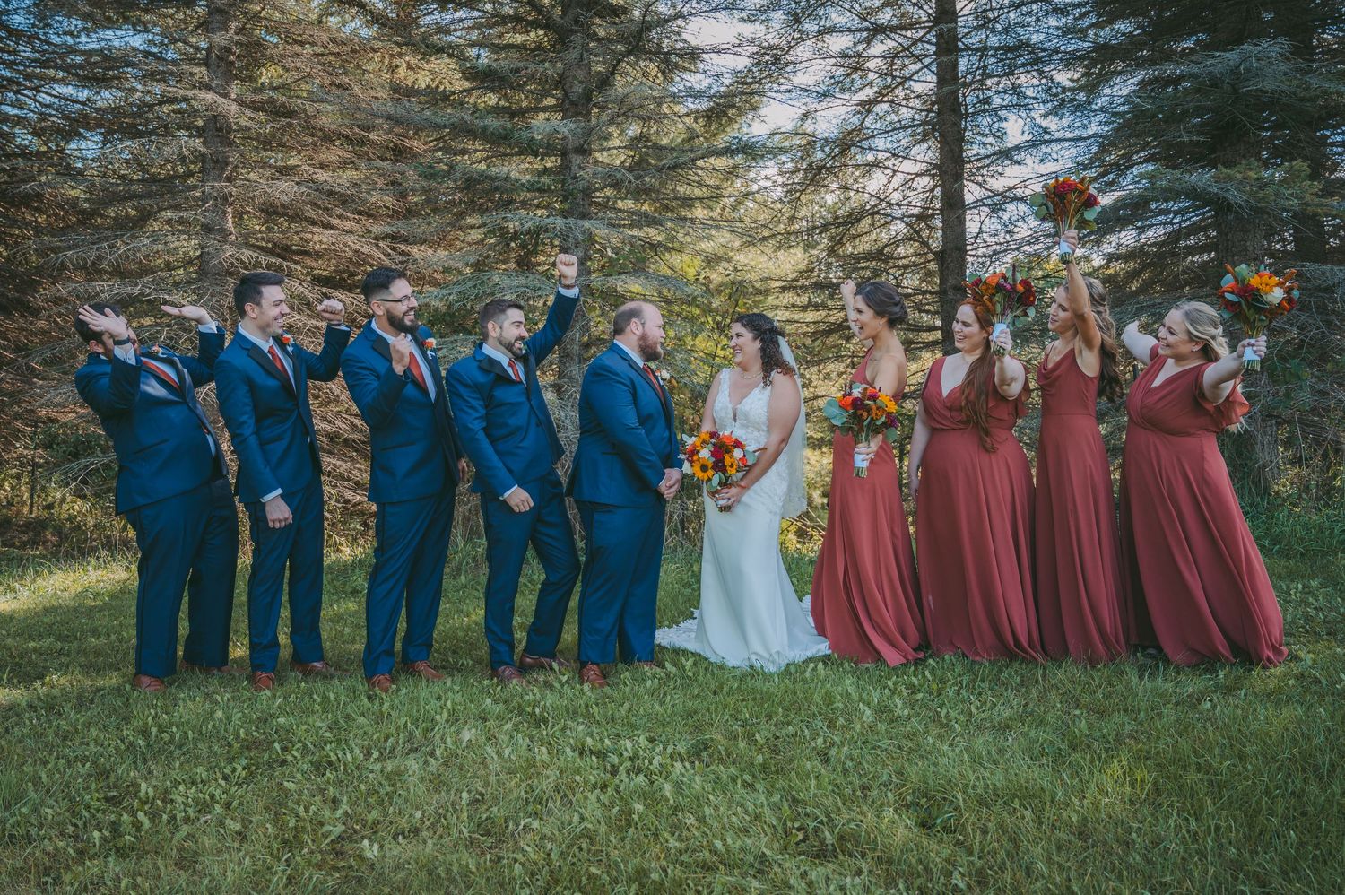 Wedding party lined up outdoors wearing navy suits and burgundy dresses against evergreen trees.