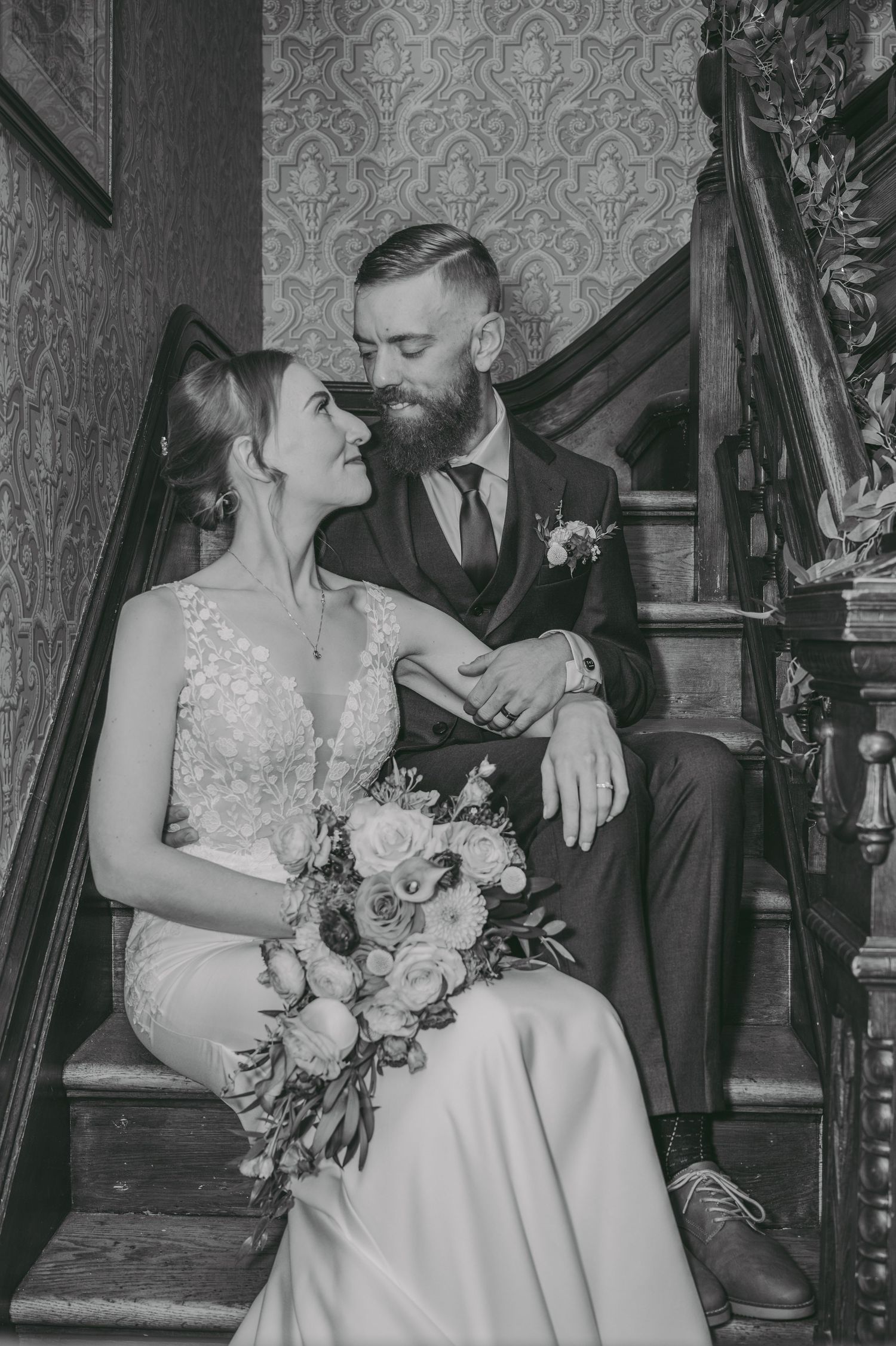 A romantic black and white wedding photo of a couple sitting on vintage stairs with ornate wallpaper.
