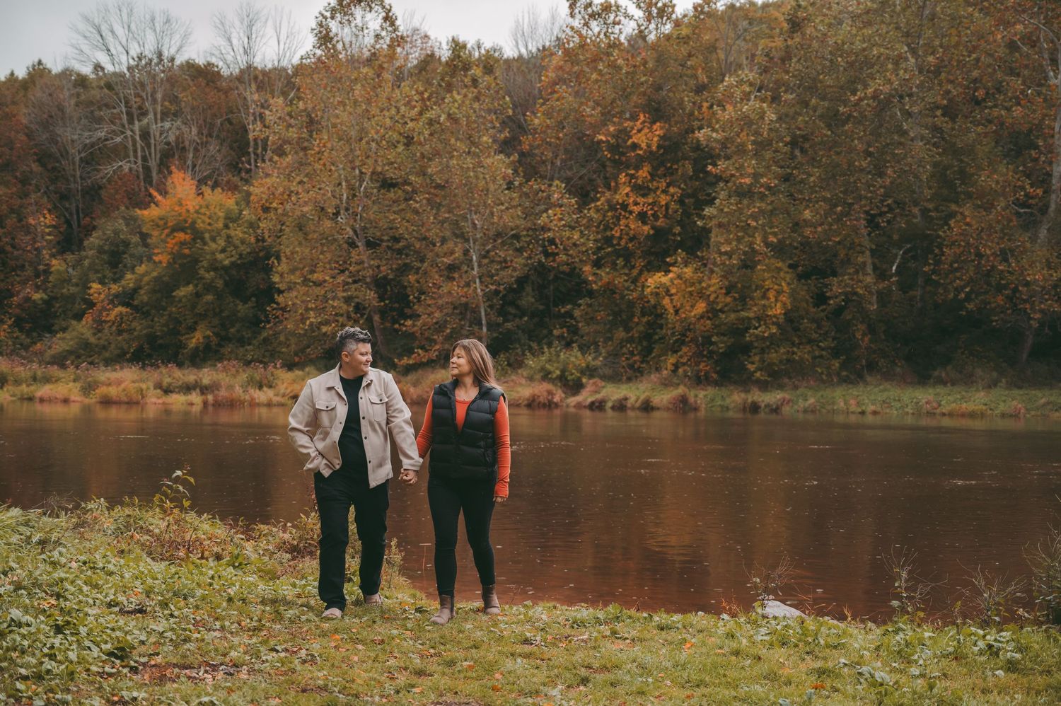 A couple standing by a serene lake surrounded by autumn trees and fallen leaves.
