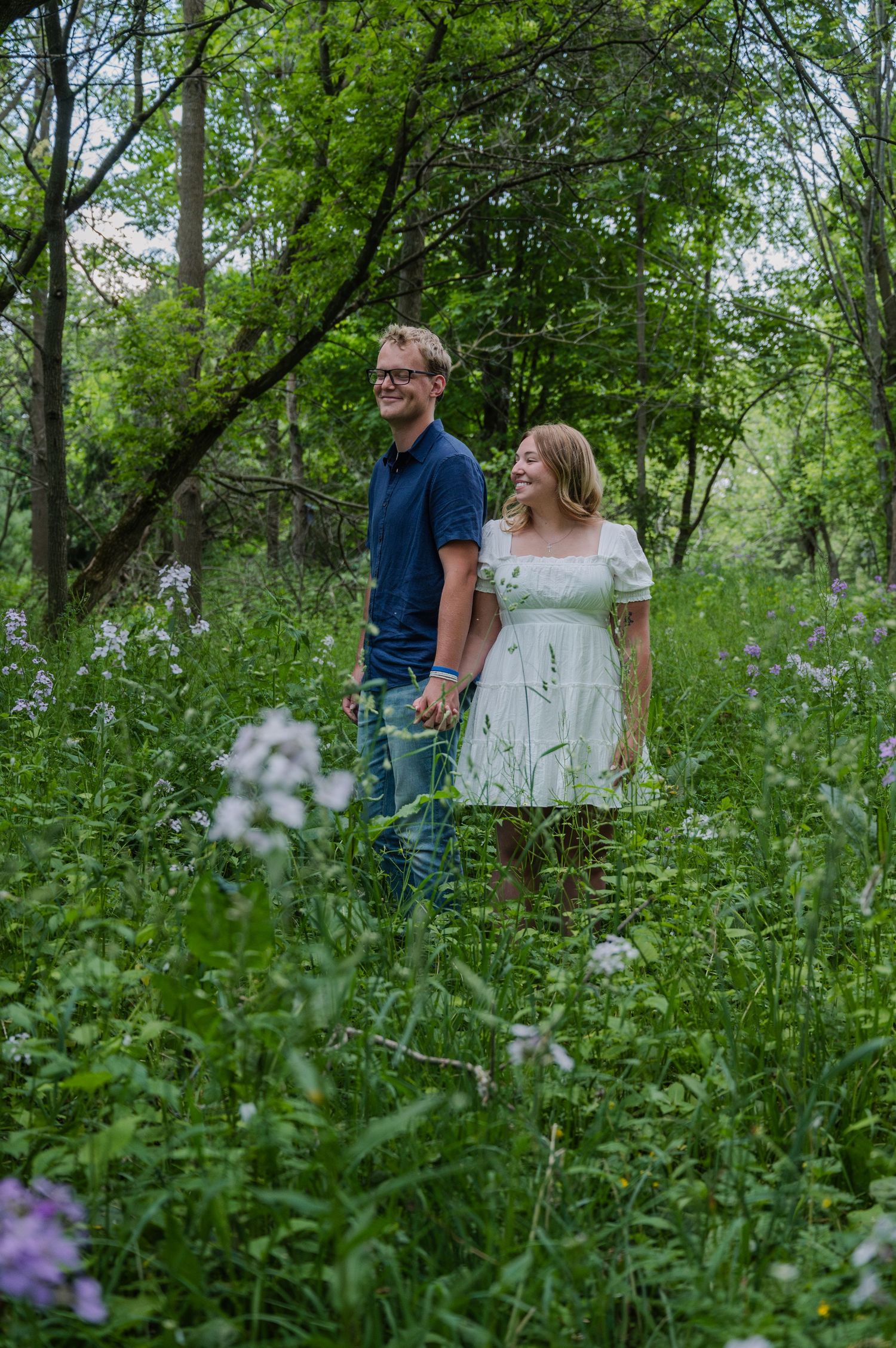 A smiling couple holds hands while standing among wildflowers and lush green trees in a romantic outdoor setting.