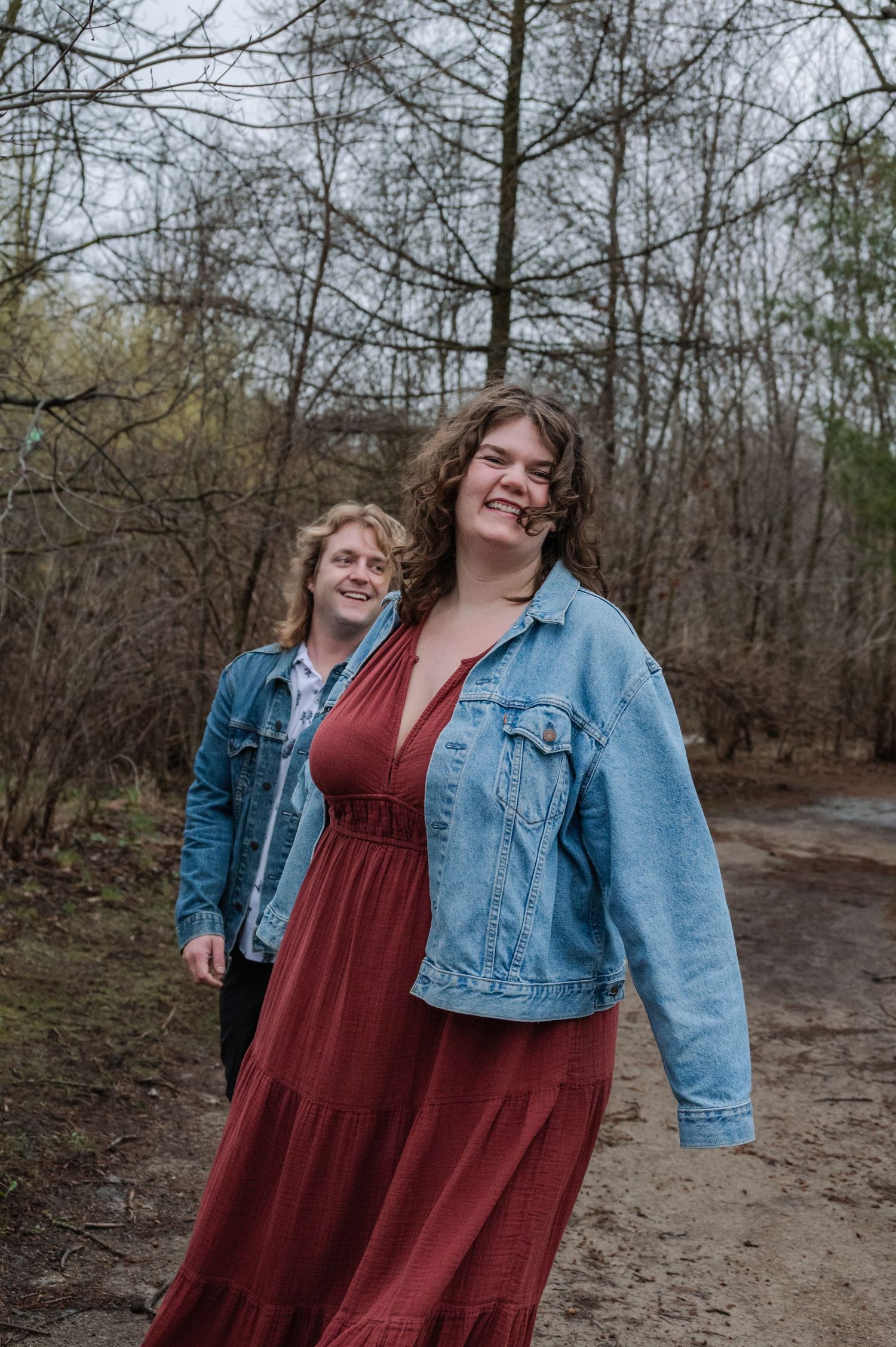 Two young women in denim jackets smile on a wooded trail, one wearing a red maxi dress in the foreground.