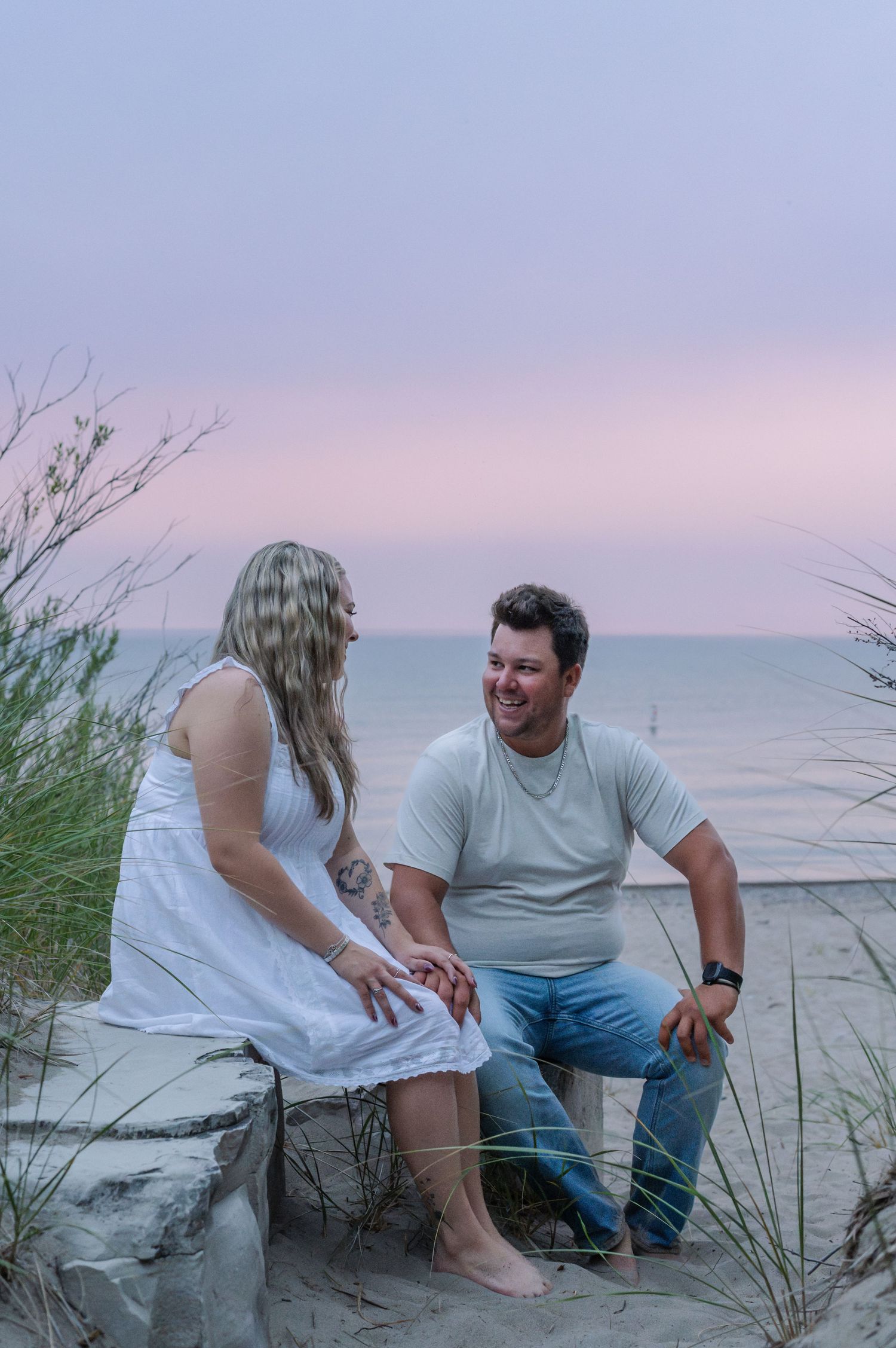 Couple sitting on beach rocks at sunset, woman in white dress and man in mint shirt smiling at each other.