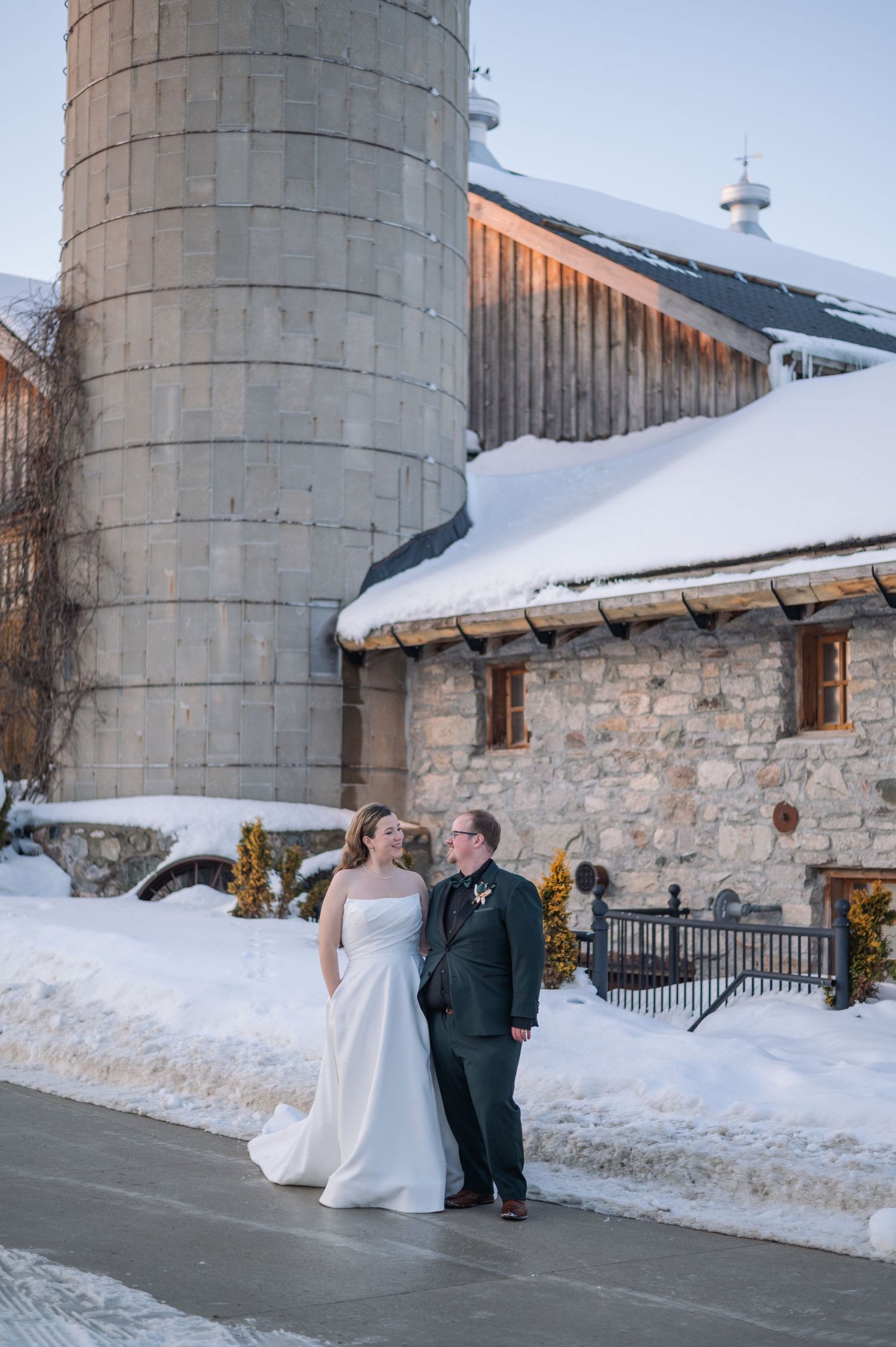 Bride and groom pose together in winter outside a snow-covered stone barn with a large silo backdrop.