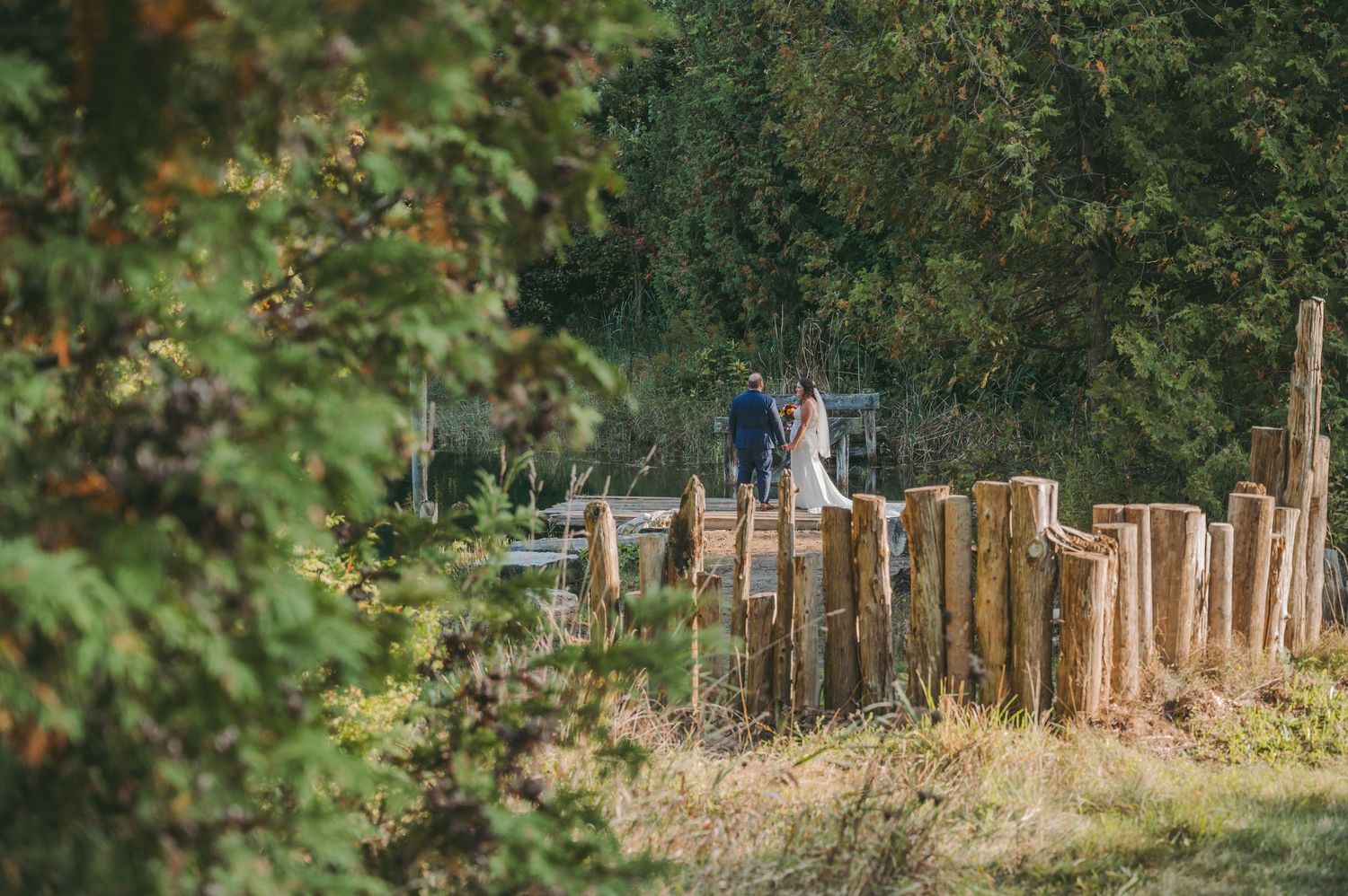 Couple in wedding attire standing by rustic wooden fence surrounded by lush green trees in outdoor nature setting.