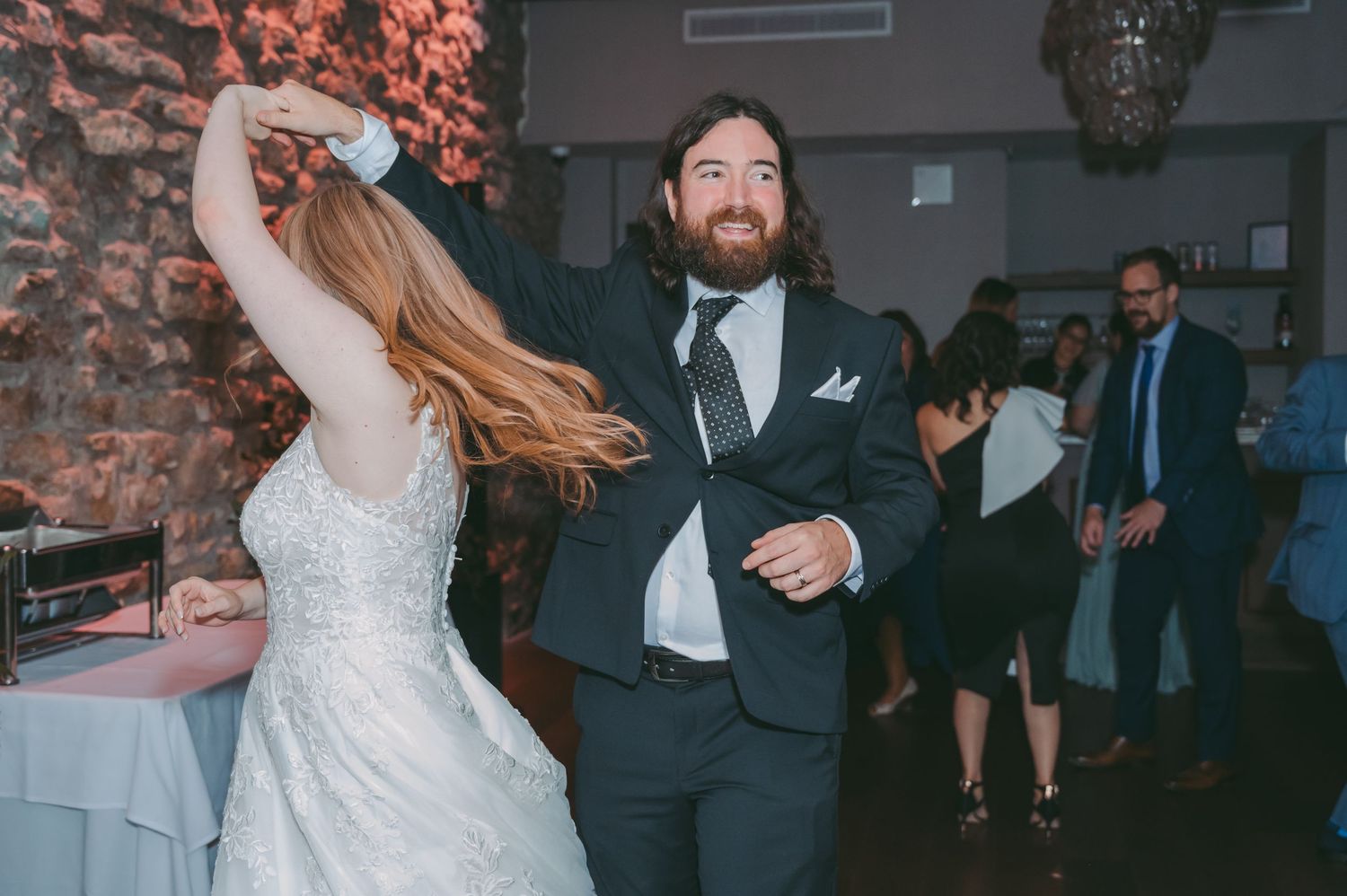 A couple dances joyfully together at their wedding reception in front of a rustic brick wall.